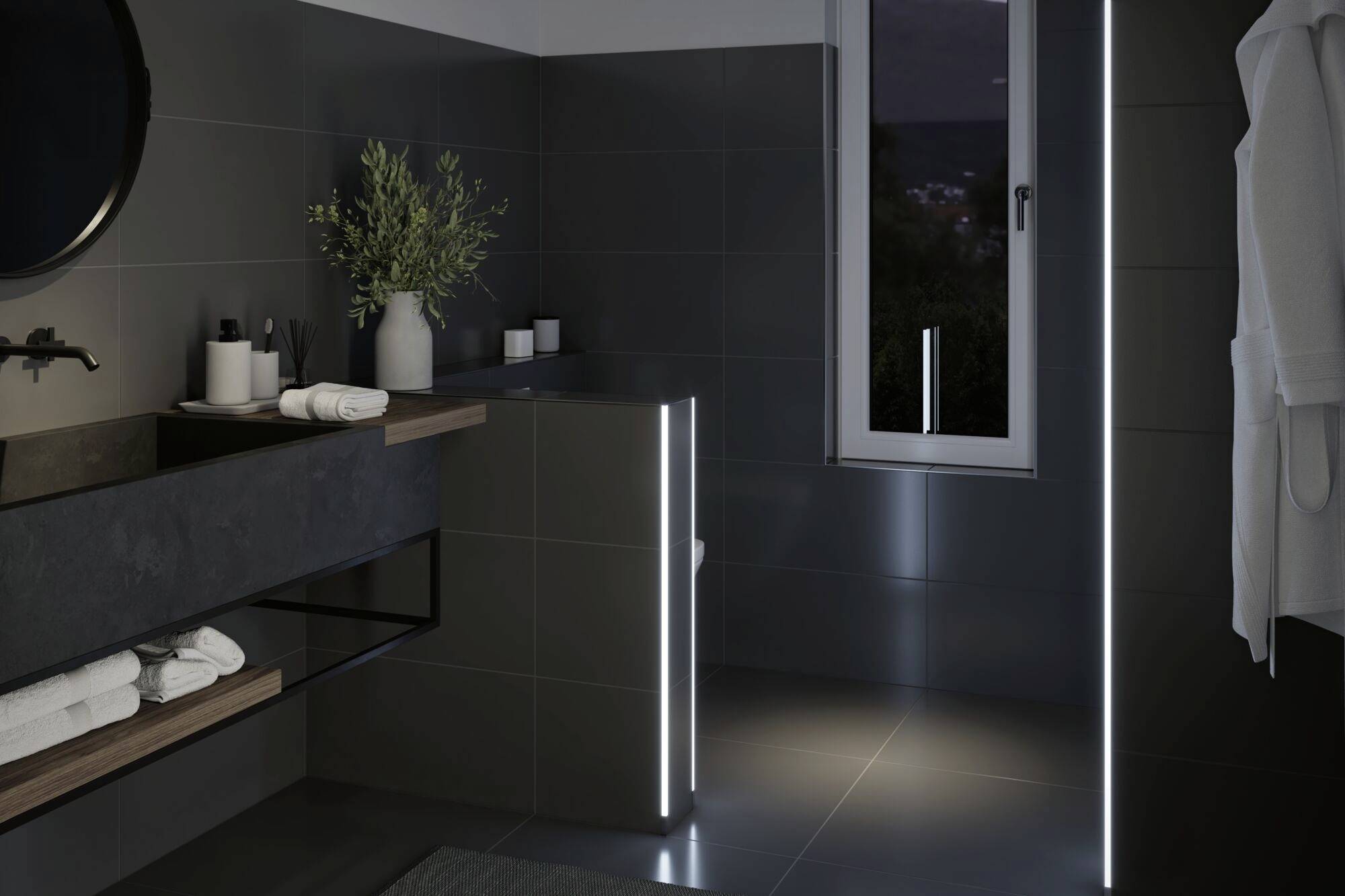 Bagno moderno con piastrelle scure, lavabo con specchio, finestra e asciugamani su una superficie di appoggio, illuminazione discreta.