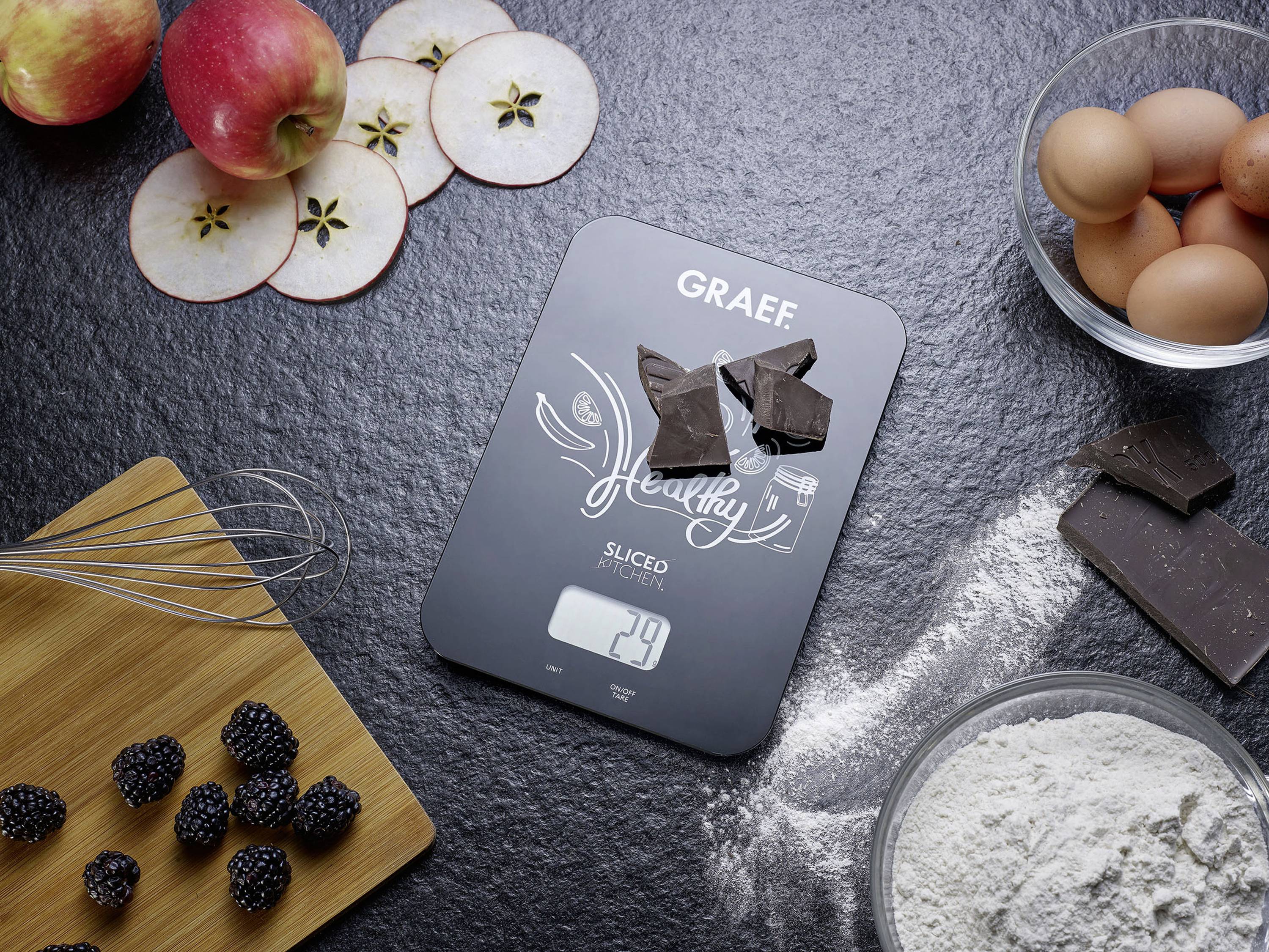 Ingredienti integrali, come mele, bacche, uova e cioccolato, circondano una bilancia digitale da cucina che mostra '29g' su uno sfondo scuro.