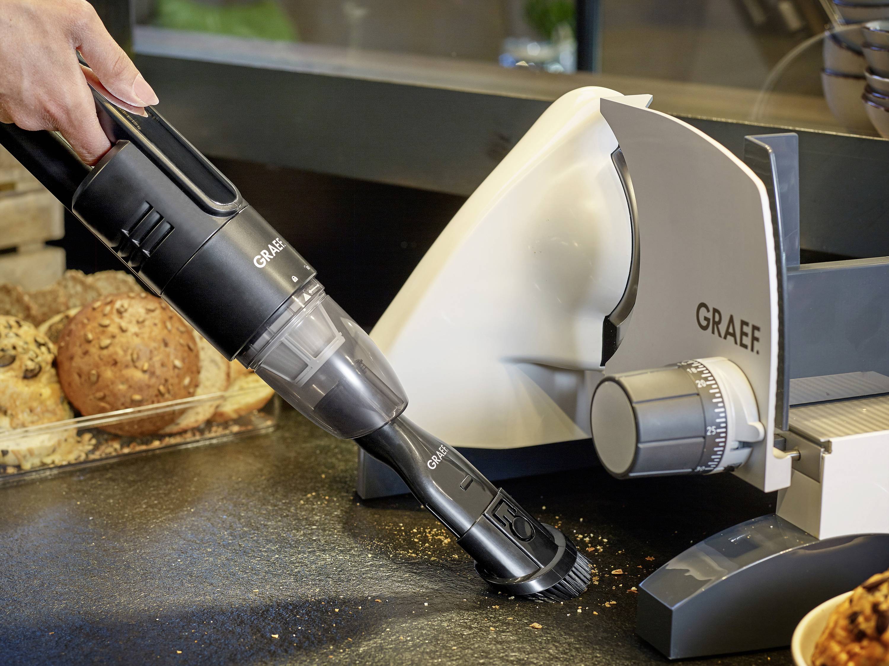 Una persona sta usando un aspirapolvere portatile per rimuovere briciole da un piano di lavoro della cucina. Accanto c'è un affettatrice per il pane.