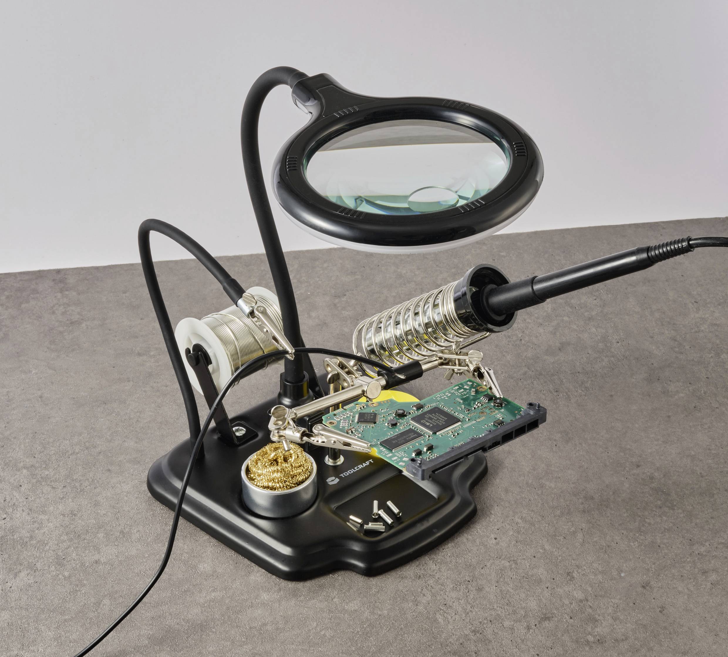 Una stazione per saldatore con una scheda elettronica; include una lente d'ingrandimento, un rocchetto di stagno e una spugna per la pulizia, posizionata su una superficie grigia.