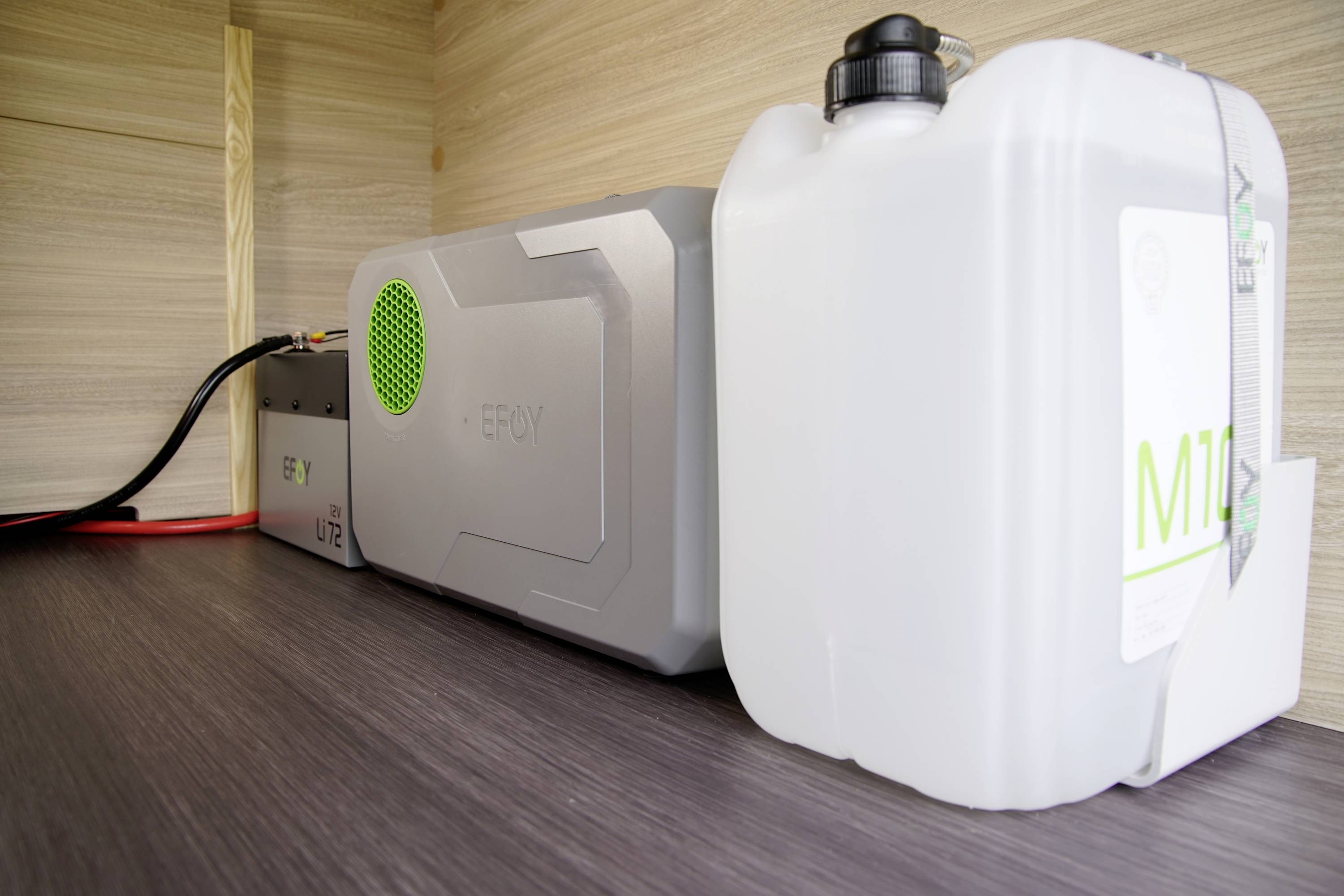 Un dispositivo di accumulo energetico grigio con ventola verde e un tanica di plastica bianca sono posizionati su una superficie di legno.