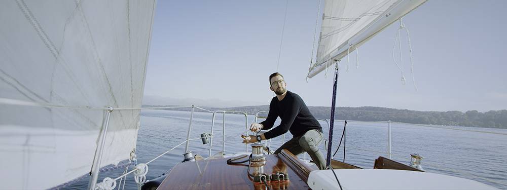 Un uomo è in piedi su una barca a vela e tira una cima. Sullo sfondo si vedono acque calme e una costa boscosa.