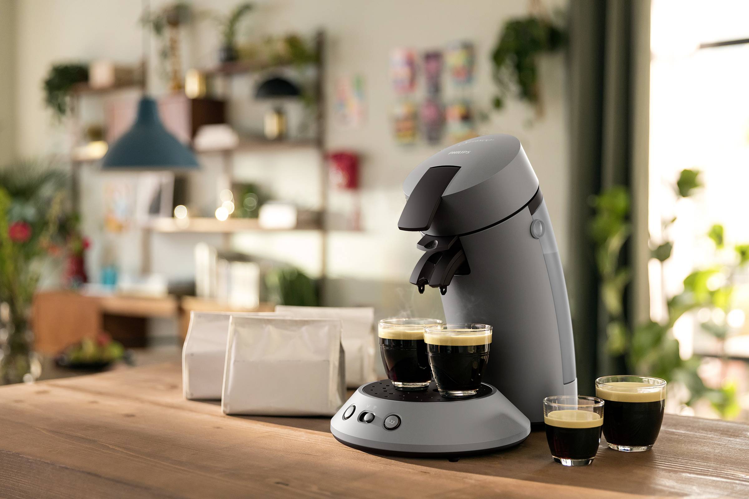 Una macchina da caffè grigia è posizionata su una tavola di legno, con quattro bicchieri di caffè accanto. Sullo sfondo sfocato di un ambiente moderno e accogliente.