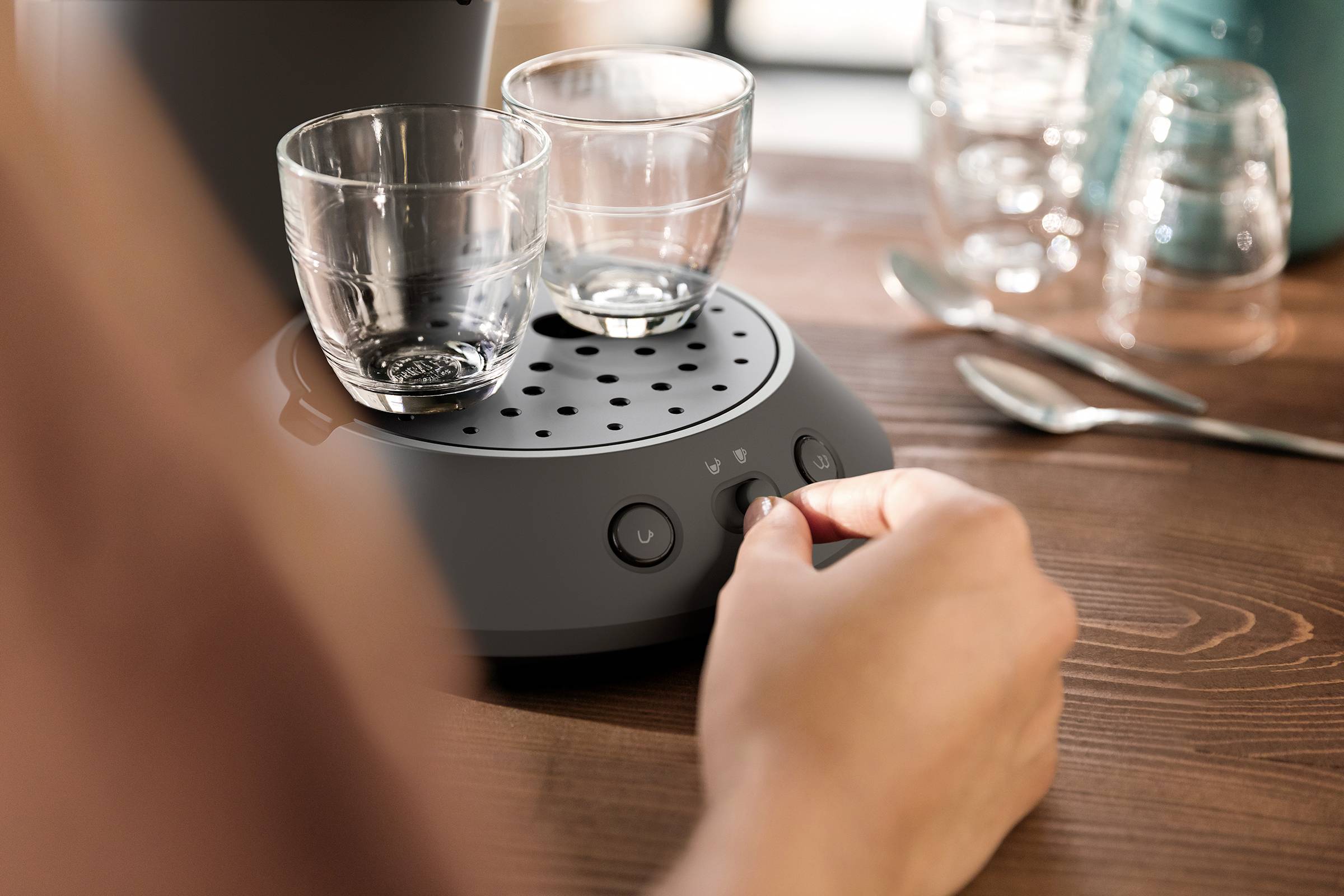 Una persona sta usando una macchina da caffè con due bicchieri sul vassoio gocciolante. Sullo sfondo ci sono dei cucchiai e altri bicchieri.
