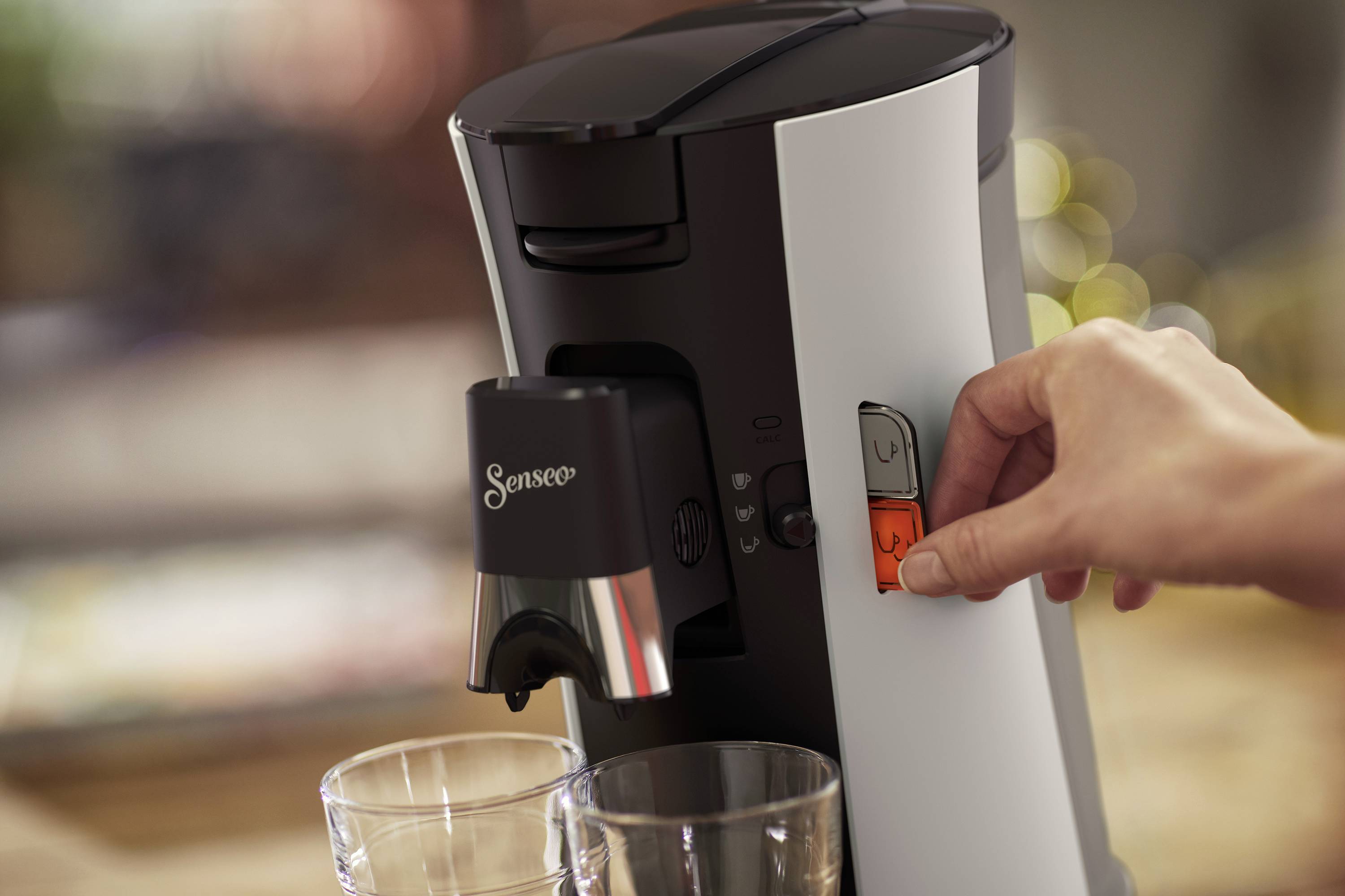 Una mano preme il pulsante di accensione di una macchina da caffè. Due bicchieri vuoti sono pronti, mentre sullo sfondo le luci appaiono sfocate.