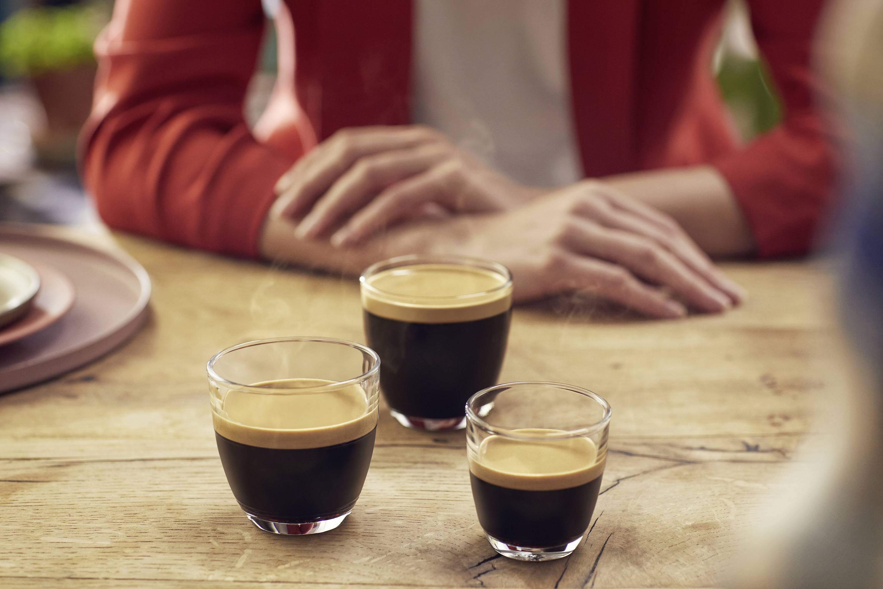 Tre tazzine da espresso su un tavolo di legno; sullo sfondo si vedono le braccia incrociate di una persona seduta.