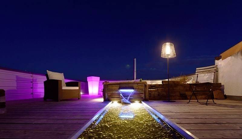 Una terrazza sul tetto elegante di notte, illuminata con colori suggestivi, arredata con mobili per sedersi e un corso d'acqua decorativo illuminato in modo atmosferico.