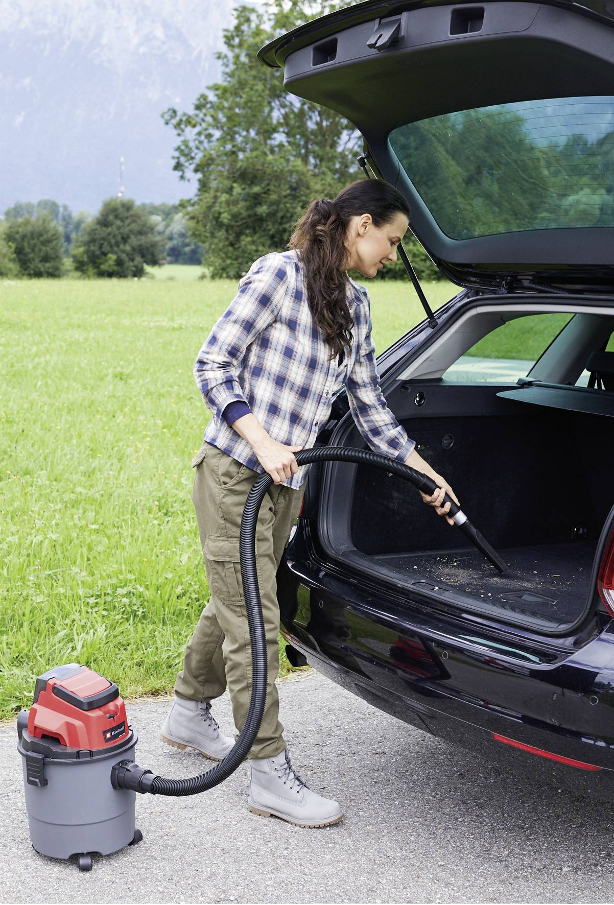 Una donna aspira l'interno del bagagliaio di un'auto con un aspirapolvere. Sullo sfondo si vede un prato verde.