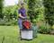 Un uomo sta triturando residui vegetali nel giardino con un cippatore rosso. Sullo sfondo si vedono piante curate e un grande sacco per le foglie.