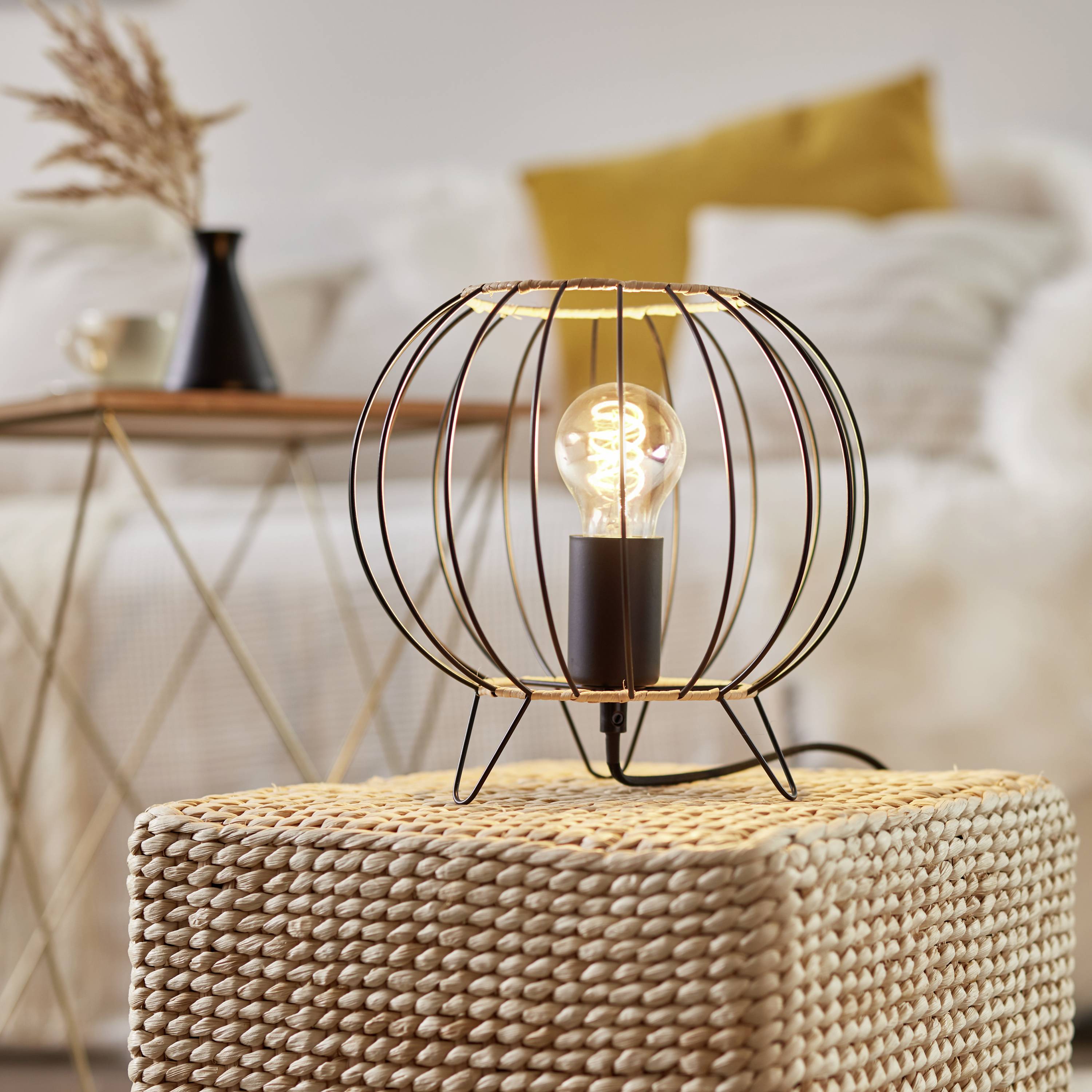 Una lampadina moderna in una gabbia metallica rotonda su uno sgabello intrecciato. Sullo sfondo un divano con cuscini e un tavolo.