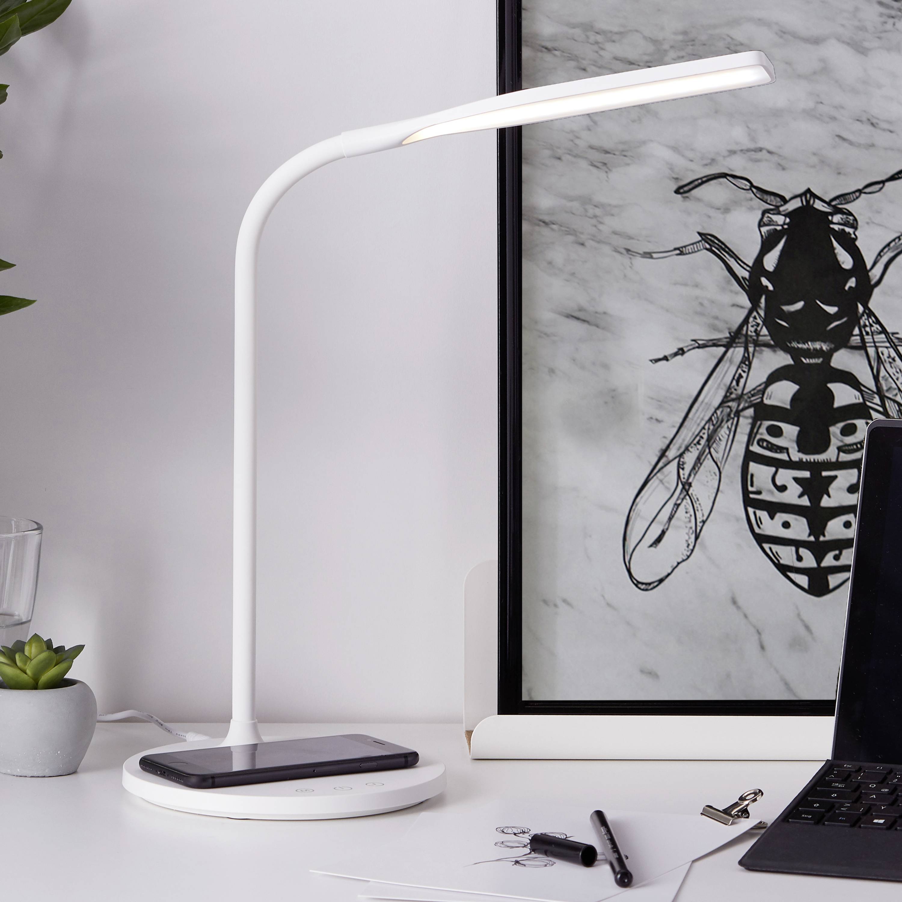 Un elegante scrittoio con una lampada che carica wirelessly un cellulare. Accanto, un'opera d'arte con un motivo geometrico di insetti sullo sfondo.