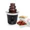 Una fontana di cioccolato con cioccolato fluente è posizionata su un tavolo. Accanto ci sono fragole, marshmallow e biscotti come accompagnamento.