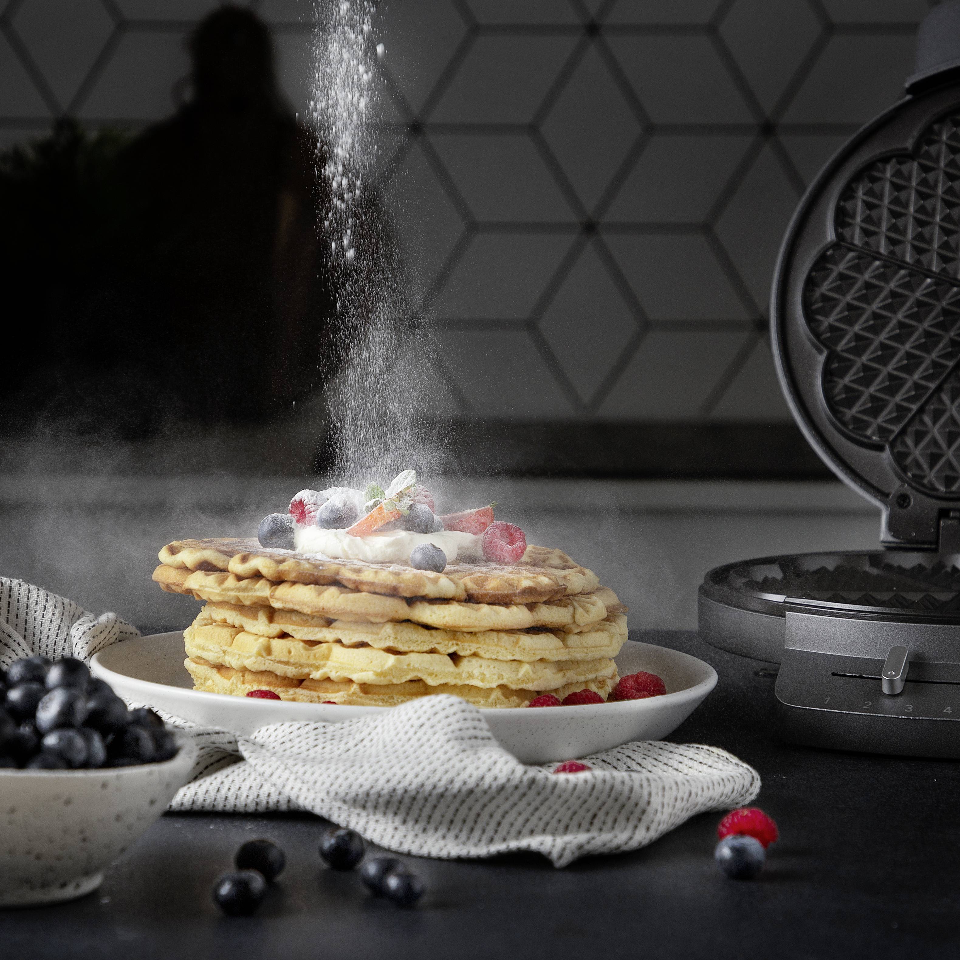 Un mucchio di pancake ricoperti di zucchero a velo, con lamponi e mirtilli su un piatto. Accanto, sulla superficie del bancone, è presente una piastra per waffles.