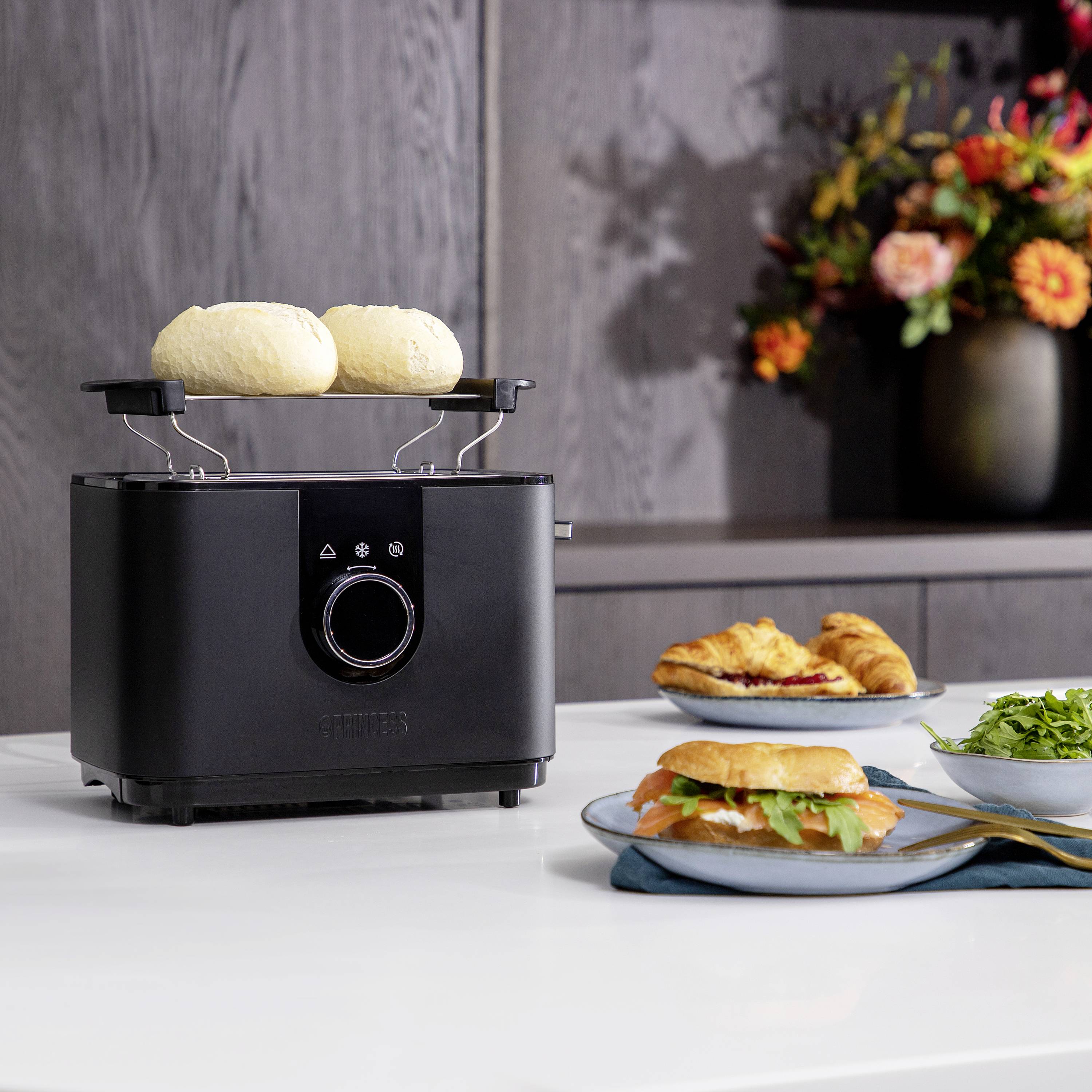 Un tostapane nero sta tostando due panini. Sullo sfondo ci sono dei fiori e sul tavolo sono posati un panino e un'insalata.