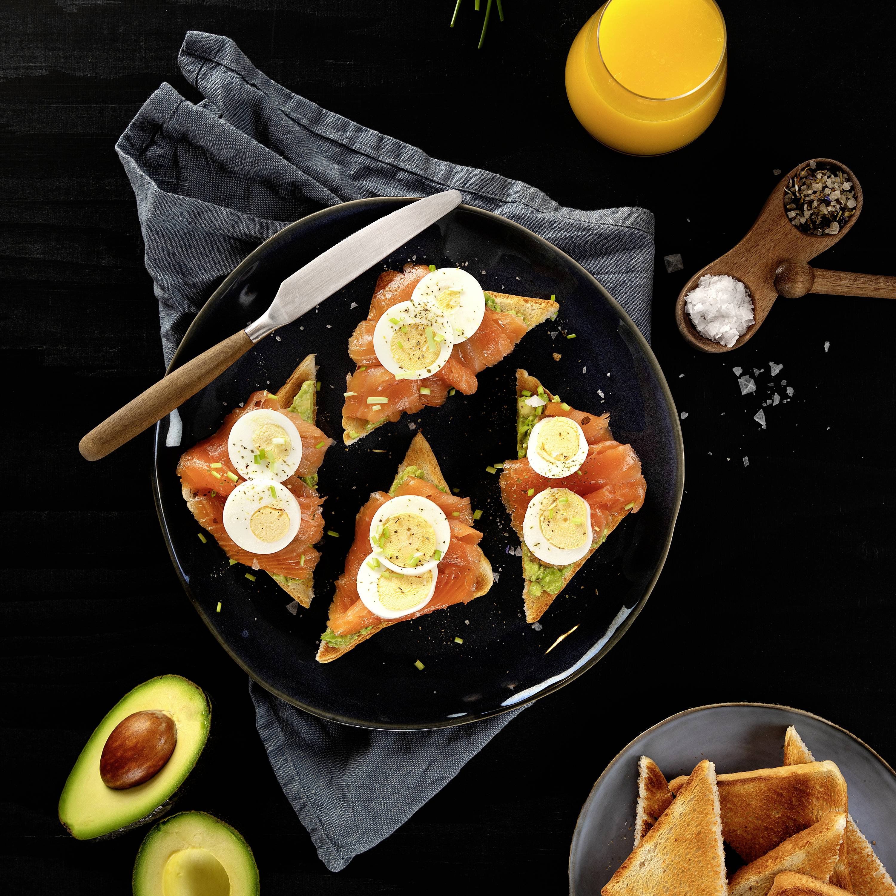 Un piatto con toast all'avocado, guarnito con salmone e fette di uova sode, accanto a un bicchiere di succo d'arancia e spargisale e pepe.