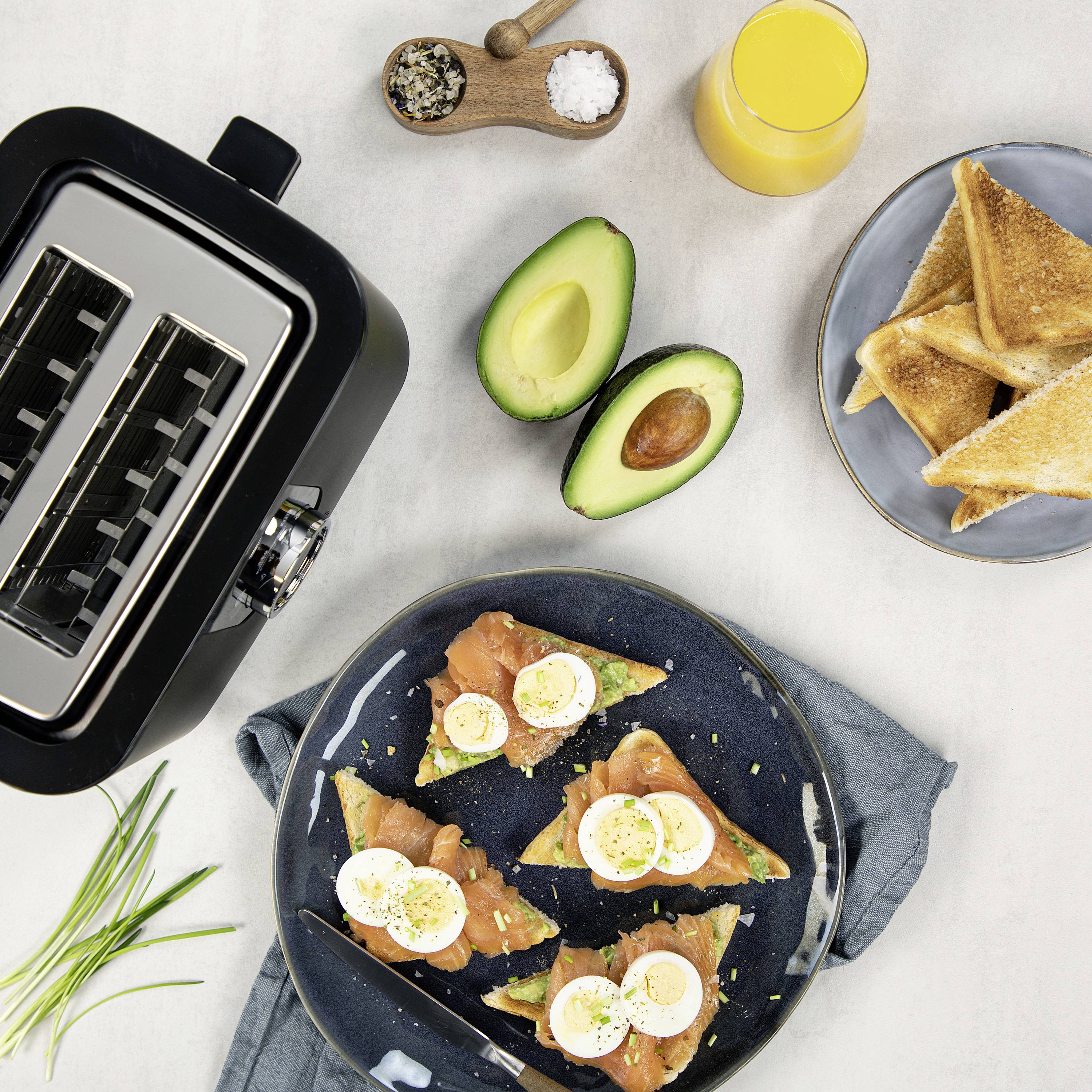 Scena di colazione con tostapane, avocado, salmone affumicato su pane e uova sode, accanto a un bicchiere di succo d'arancia e toast gratinati.