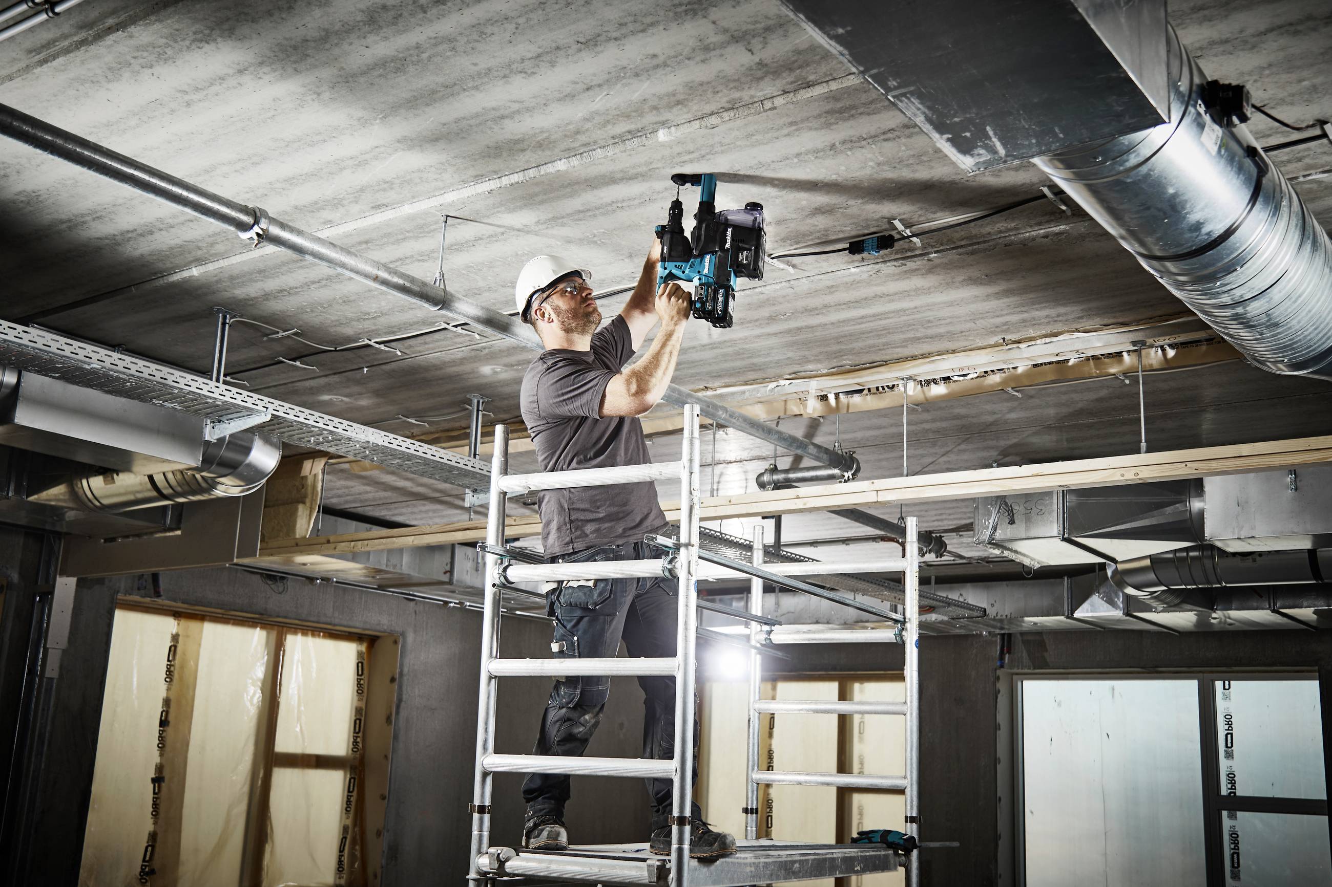 Un muratore è in piedi su un ponteggio e sta trapanando il soffitto di un edificio in costruzione. Indossa un casco di protezione e utilizza un trapano elettrico.