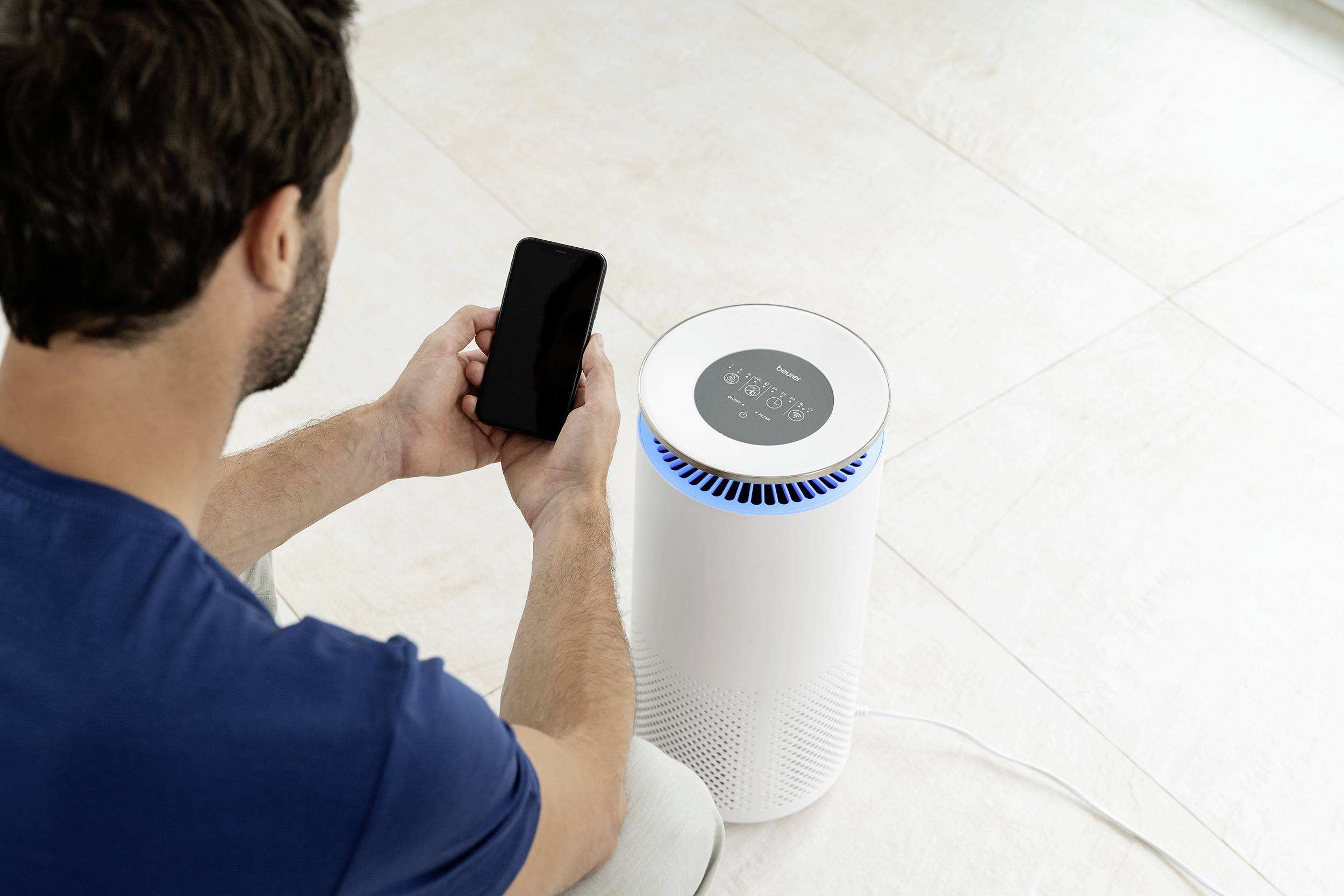 Un uomo controlla un purificatore d'aria posto sul pavimento utilizzando il proprio smartphone. Il display mostra informazioni sulla qualità dell'aria.