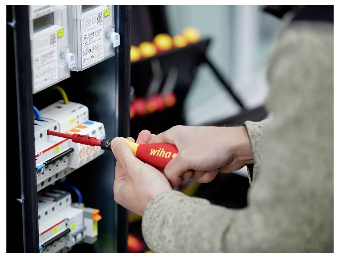 Una persona che utilizza un cacciavite rosso e giallo per regolare i collegamenti elettrici all'interno di un pannello di quadro elettrico.