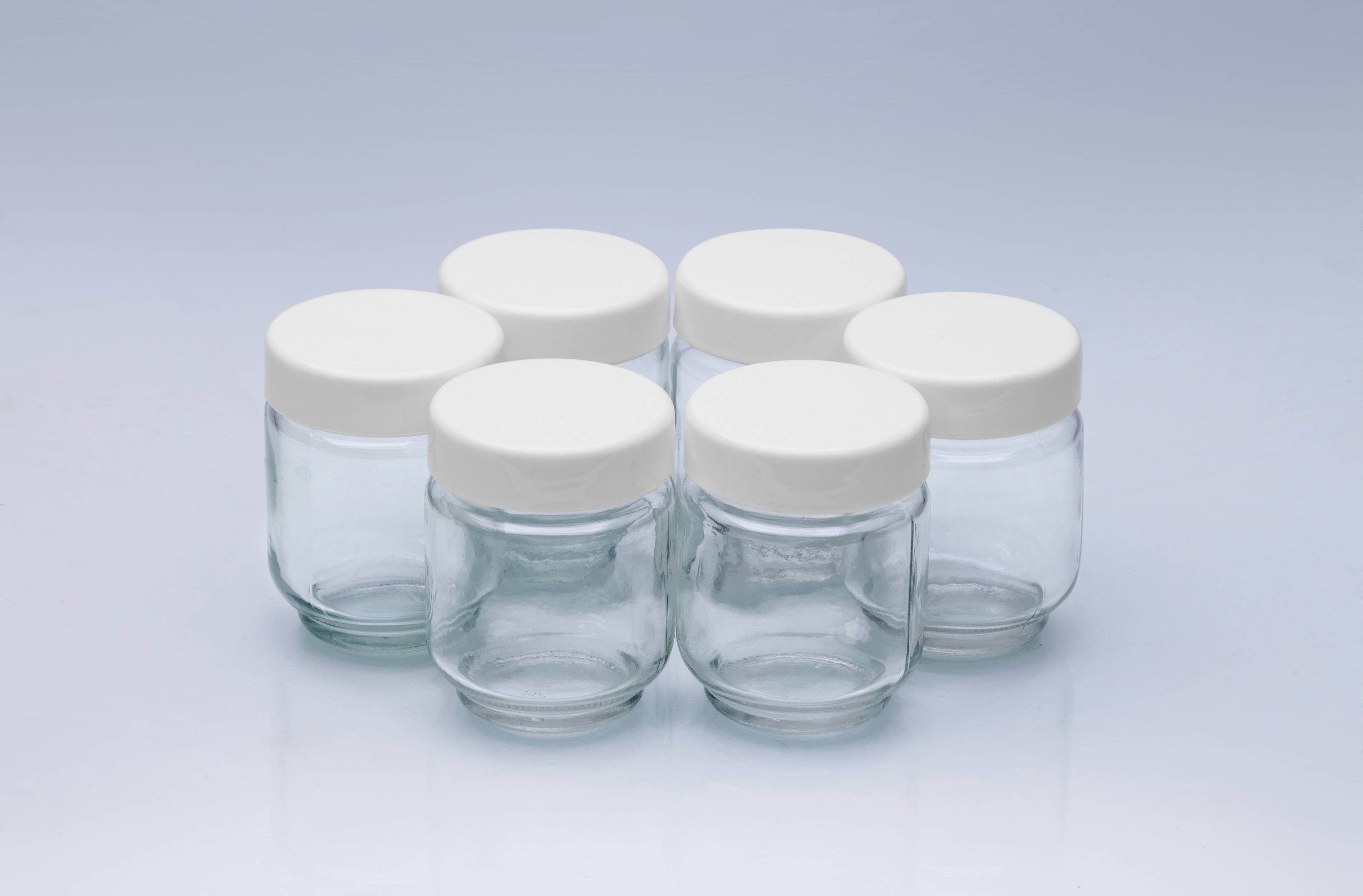 Sei barattoli di vetro vuoti con coperchi bianchi sono disposti in cerchio su uno sfondo azzurro chiaro.