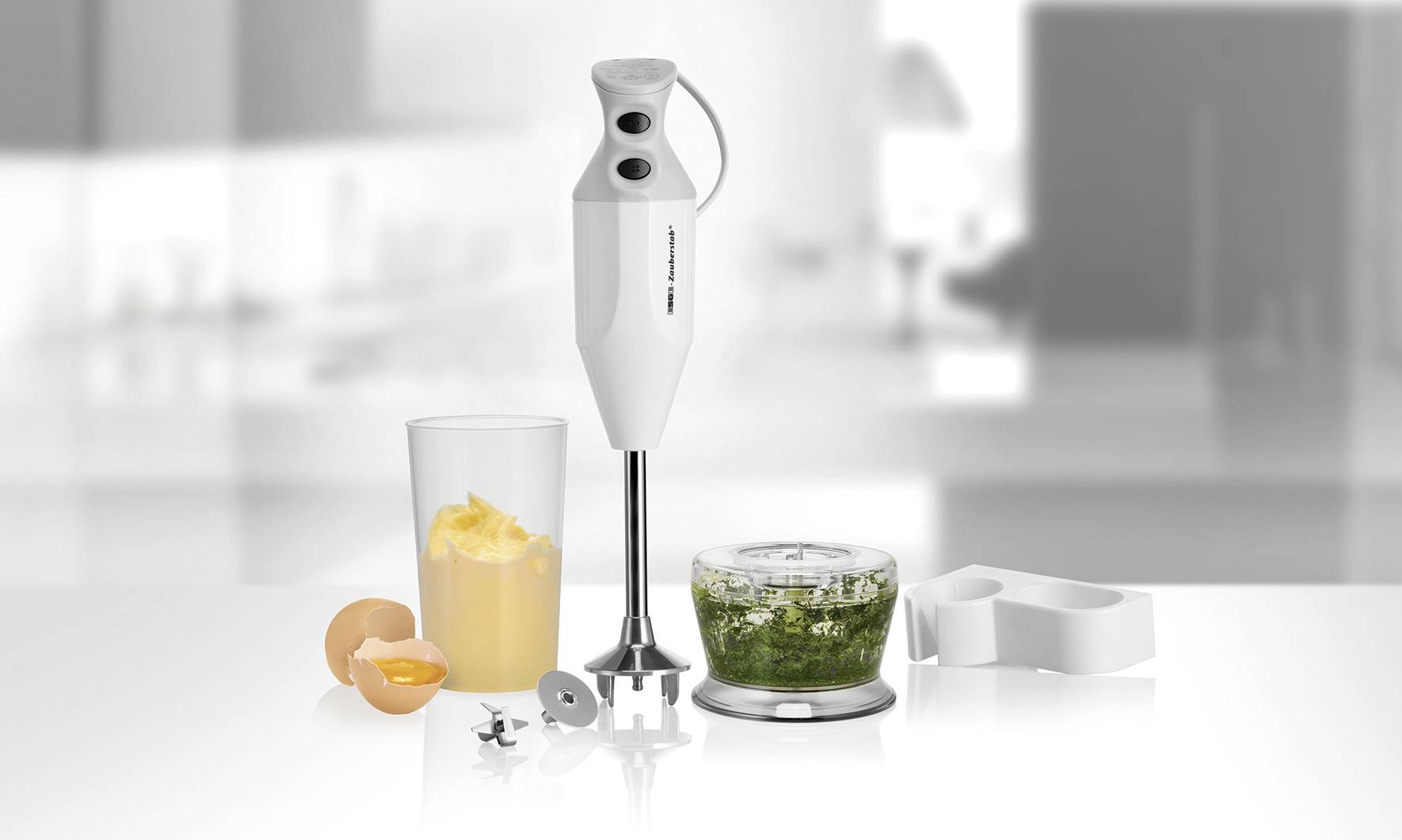 Un frullatore a immersione con diversi accessori, un bicchiere con succo giallo e un contenitore con purè verde, su un piano da lavoro in cucina.