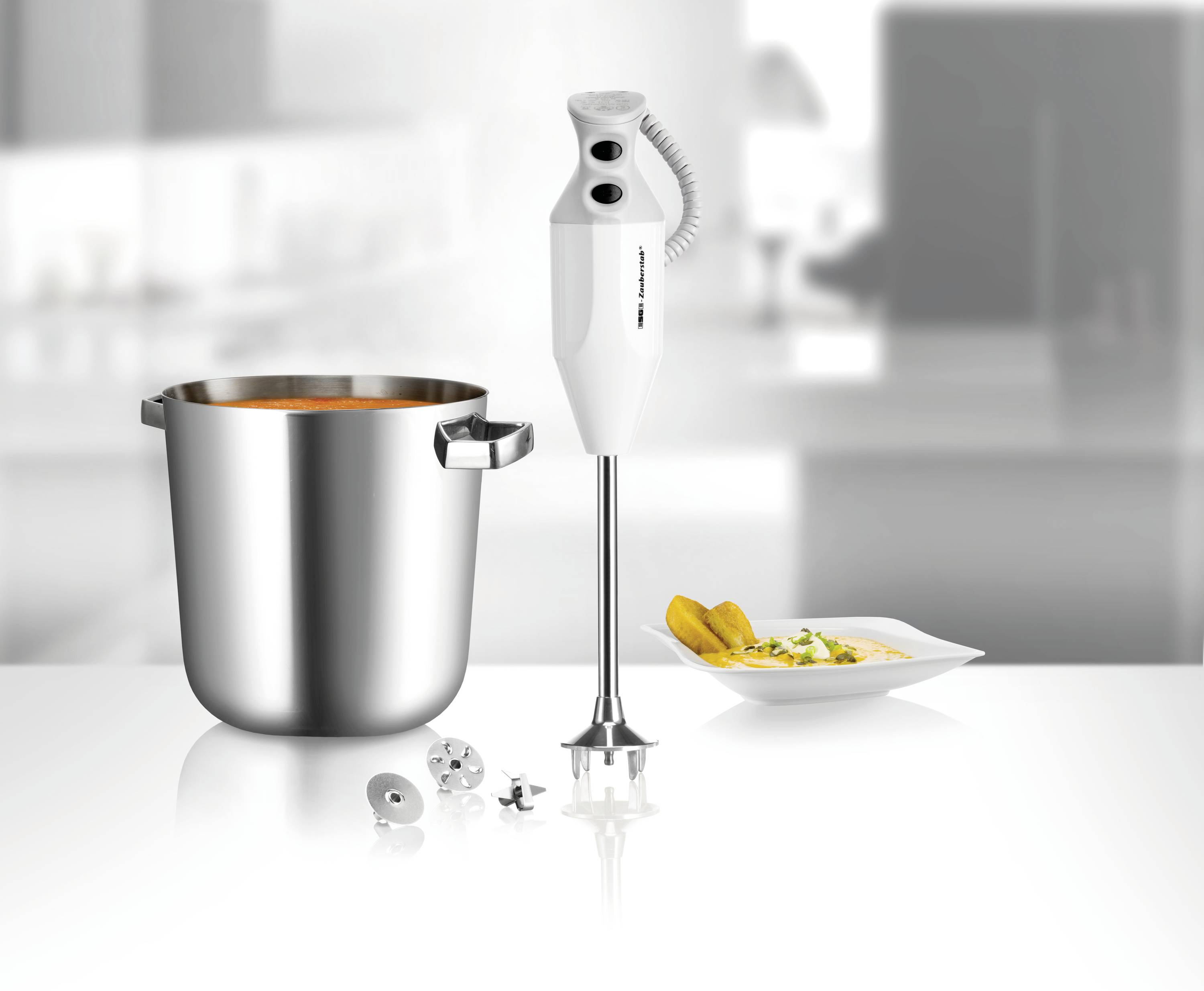 Un frullatore a immersione accanto a una pentola di zuppa e un piatto con un piatto decorato. Utensili da cucina in una cucina moderna.