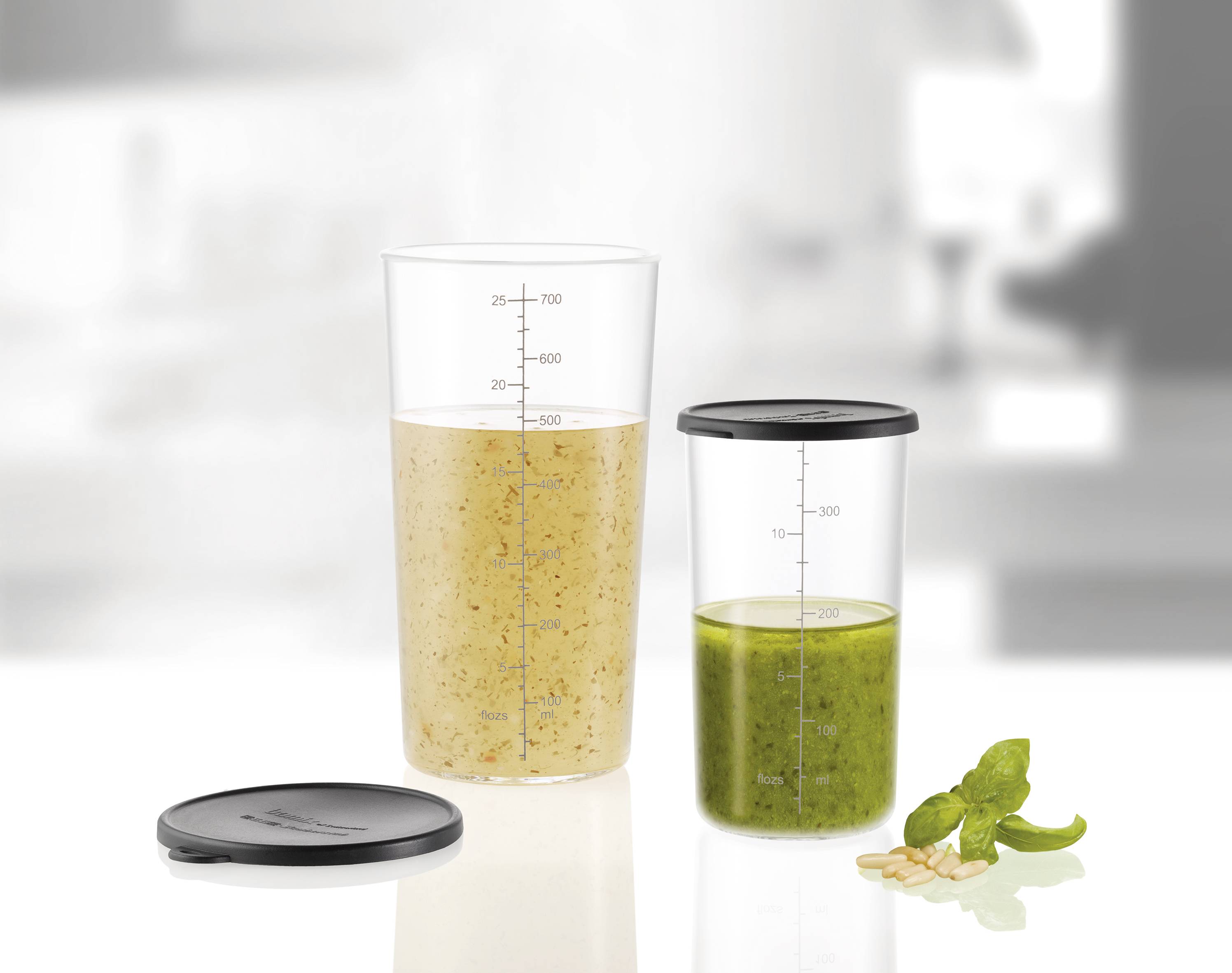 Due misurini trasparenti, quello di sinistra riempito con un liquido marrone, quello di destra con un liquido verde. Accanto un coperchio removibile, foglie di basilico e pinoli.
