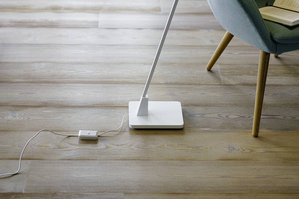 Una moderna lampada da tavolo bianca dal design sottile è posizionata su un pavimento in legno accanto a una poltrona blu.