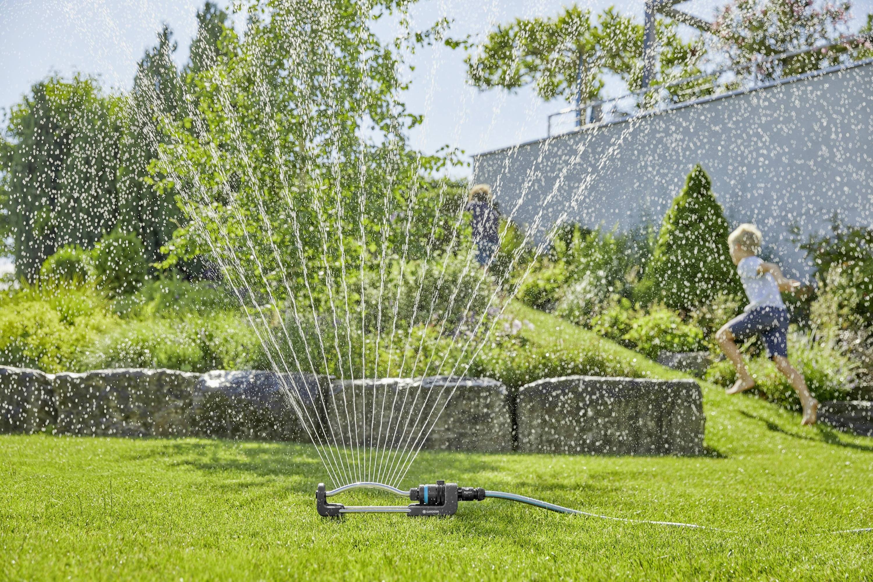 Un irrigatore spruzza acqua su un prato verde in una giornata soleggiata. Sullo sfondo, due bambini corrono attraverso lo spruzzo d'acqua.