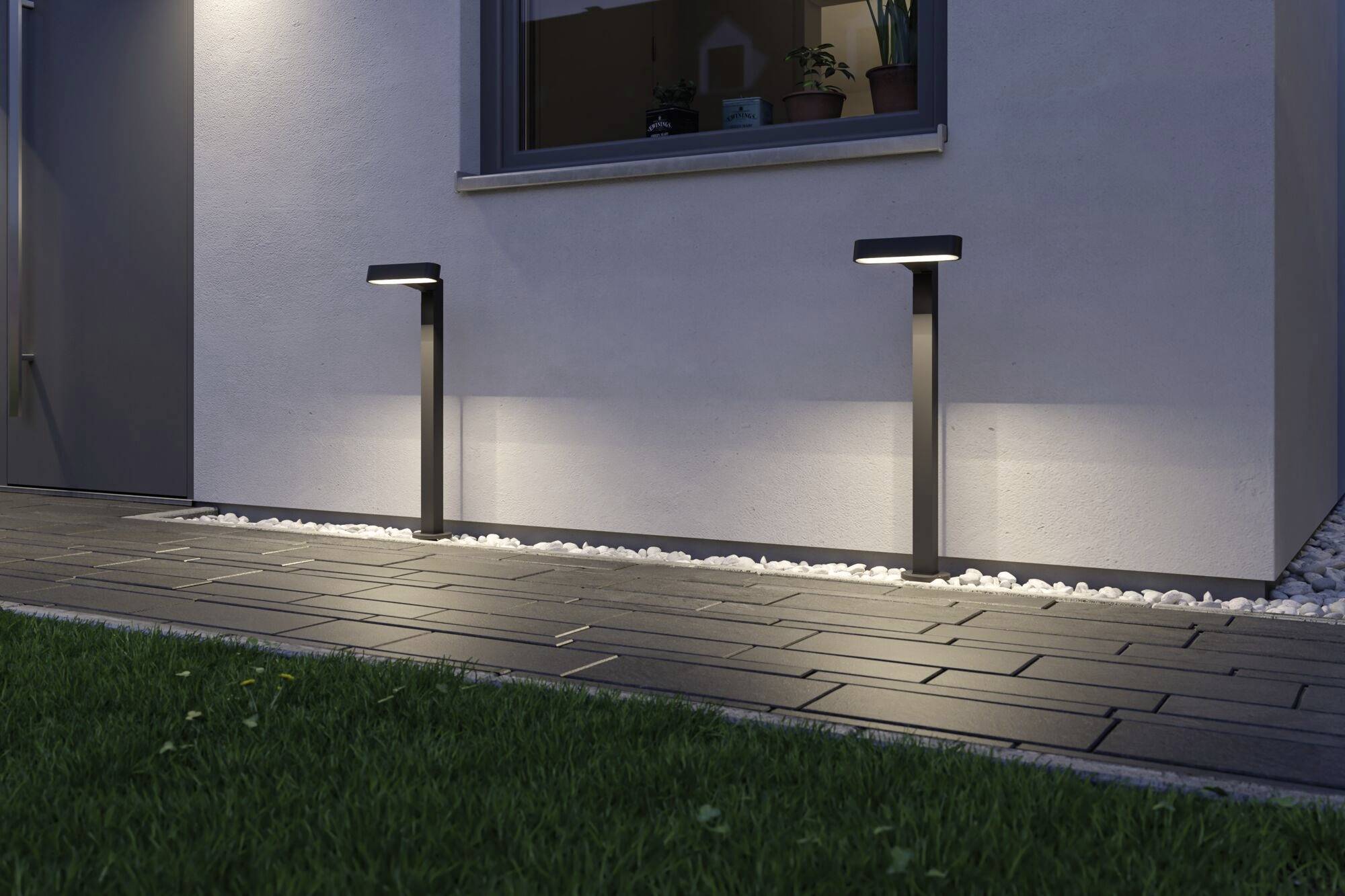 Due moderne luci esterne illuminano un percorso lastricato davanti a una casa al crepuscolo, con erba in primo piano e una finestra sullo sfondo.