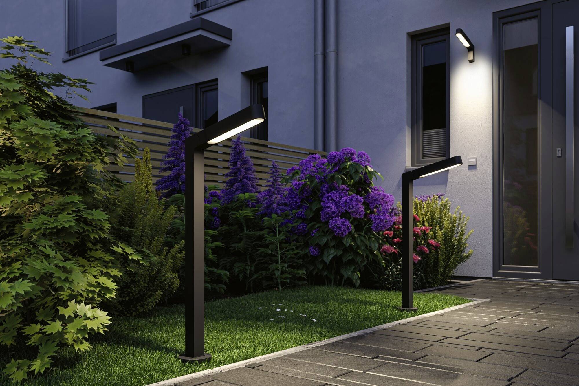 Sentiero del giardino con lampade moderne ai lati, accanto a un edificio, circondato da piante e cespugli fioriti in un'atmosfera serale.