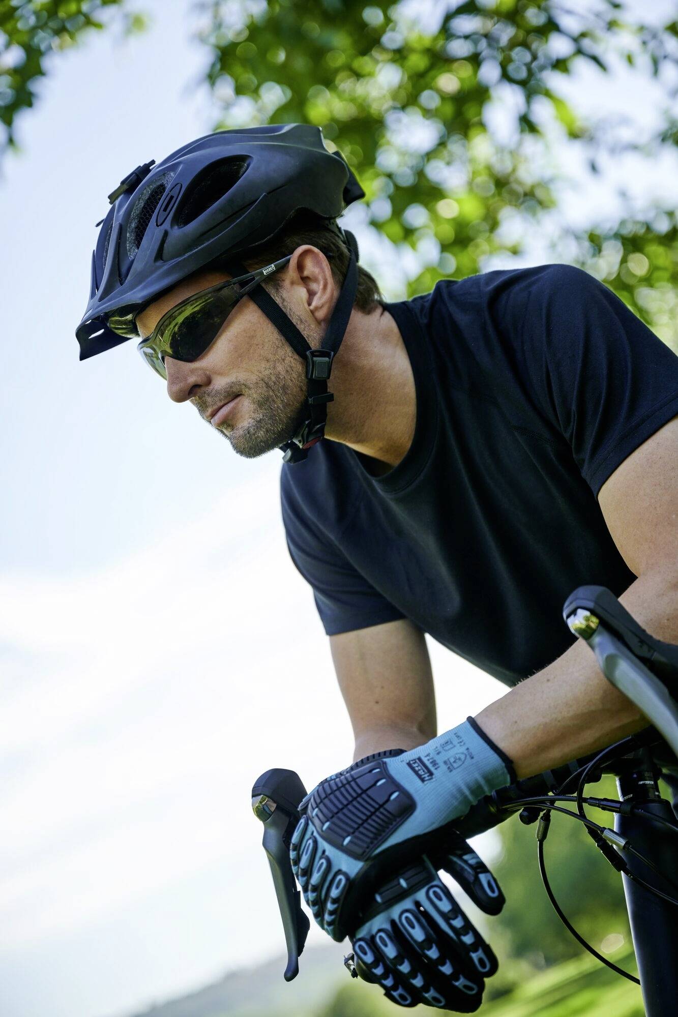 Un uomo con casco e occhiali da sole pedala in una paesaggio verde, con un cielo chiaro. Indossa una maglietta nera e guanti.