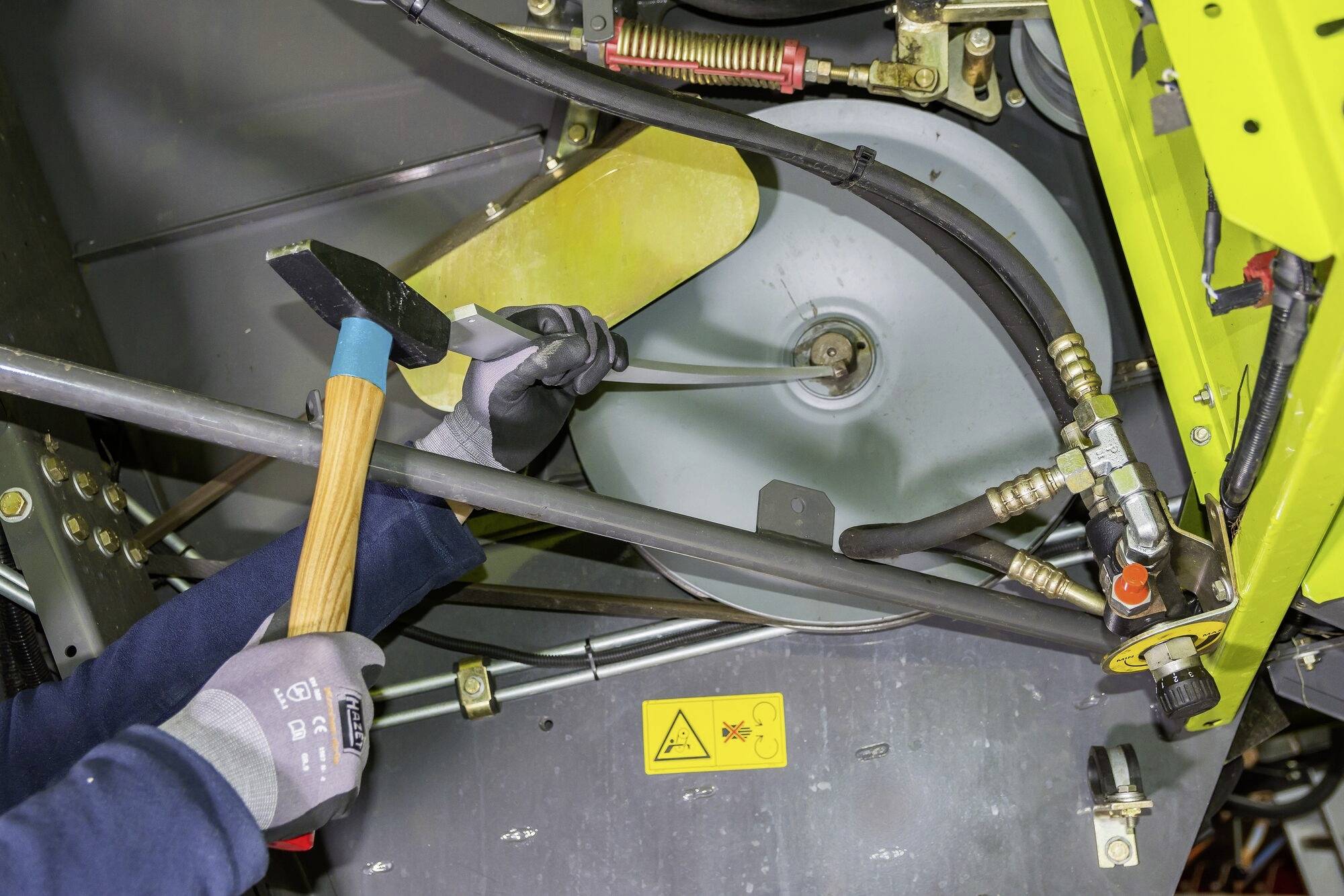 Una persona con i guanti sta lavorando su una macchina con un martello. Adesivi di sicurezza e cavi sono visibili.