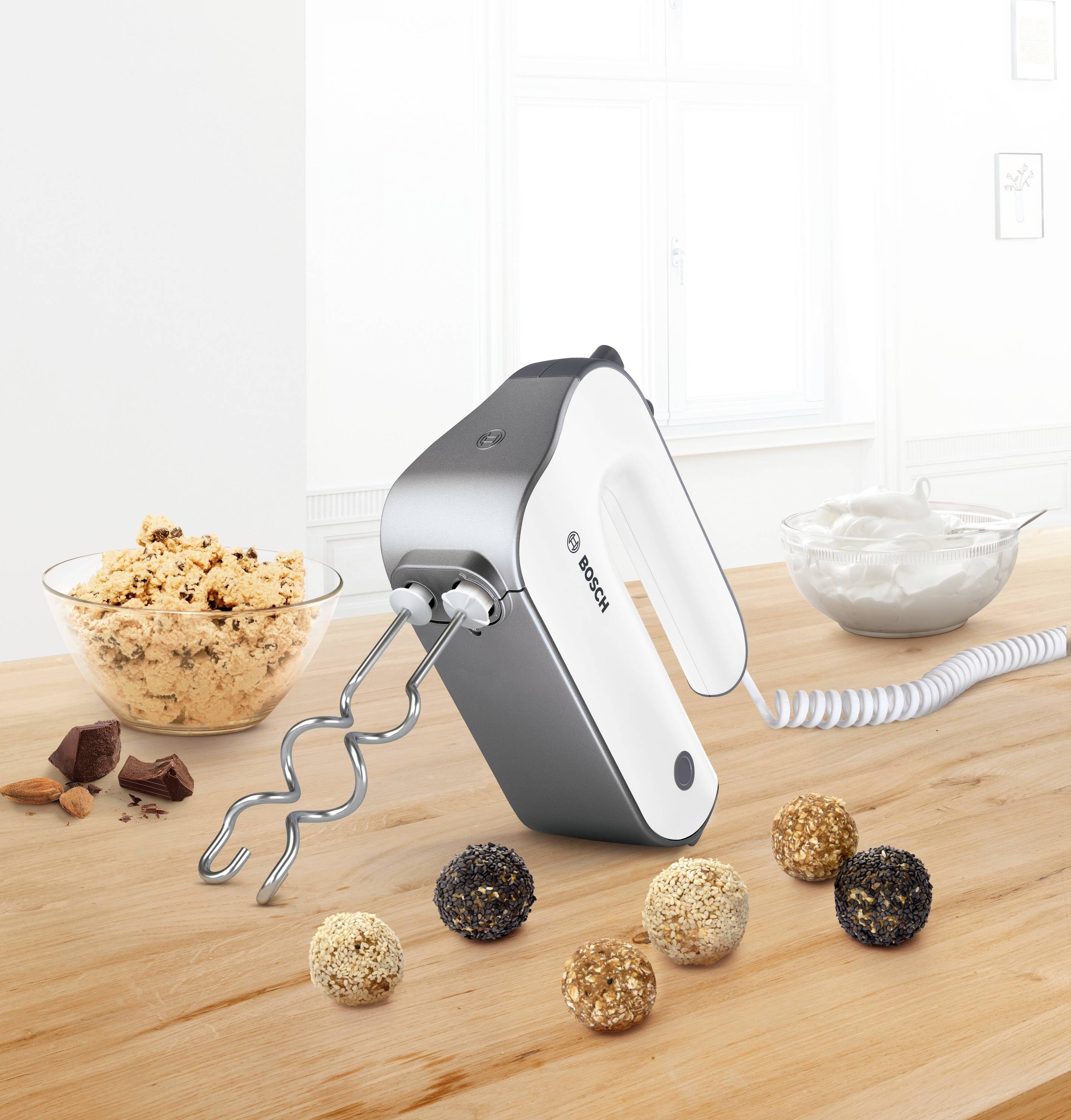 Un mixer a mano su un tavolo di legno con due ganci per impastare. Sullo sfondo ci sono ciotole con impasto per biscotti, panna e pezzetti di cioccolato.