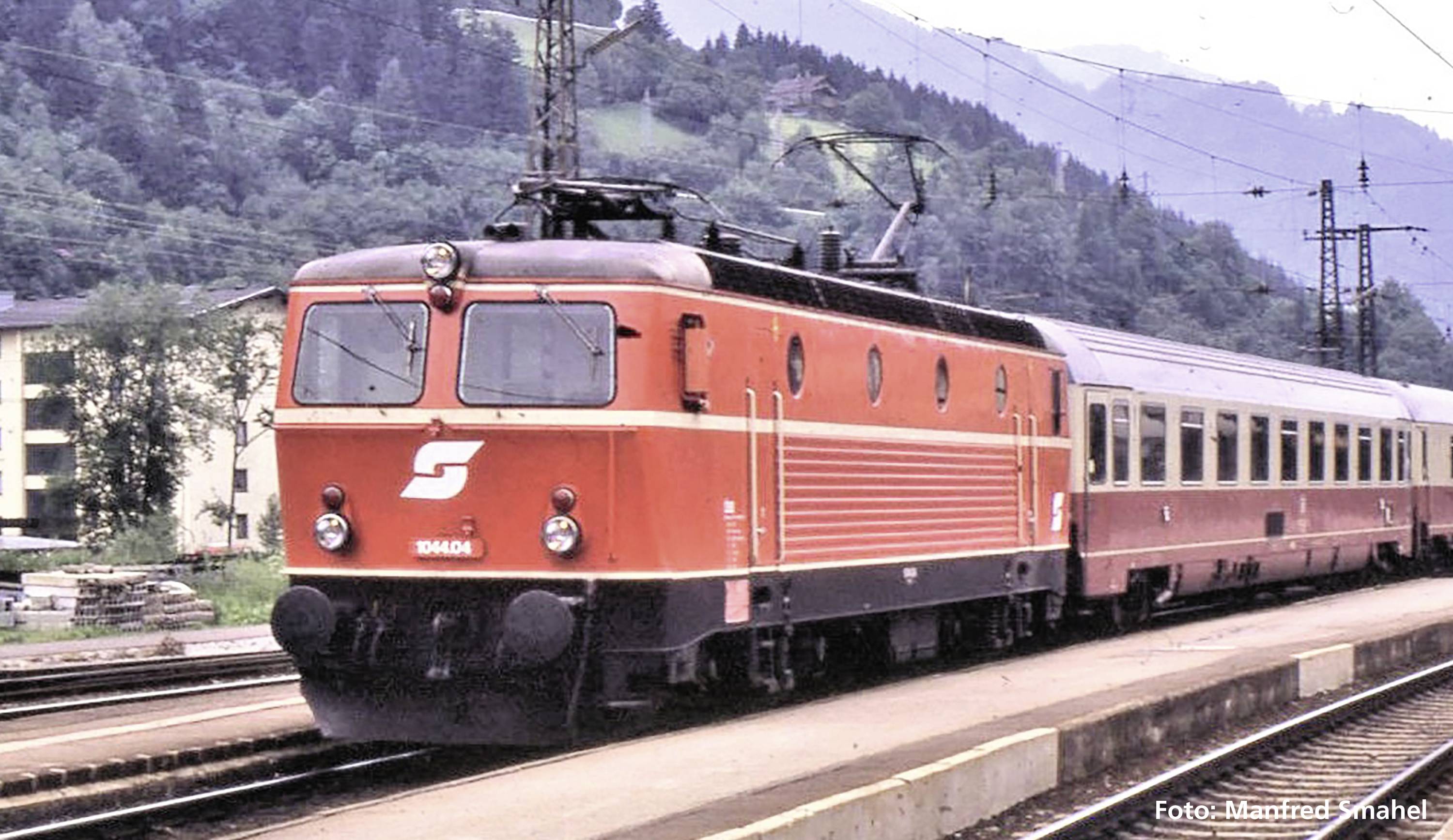 Un treno rosso con la scritta 'S' attraversa una stazione ferroviaria di campagna, con montagne e boschi sullo sfondo.