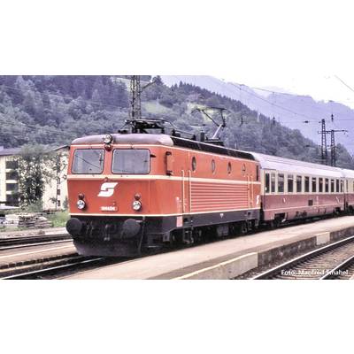 Un treno rosso con la scritta 'S' attraversa una stazione ferroviaria di campagna, con montagne e boschi sullo sfondo.