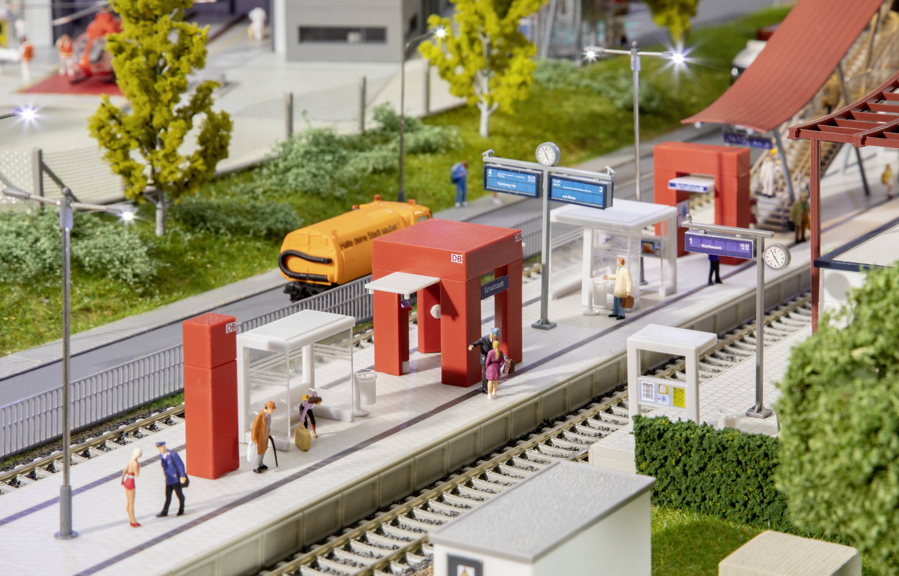 Modellino in miniatura di una stazione ferroviaria con treni, figurine e paesaggio dettagliato. Persone camminano sul marciapiede, alberi e edifici sullo sfondo.
