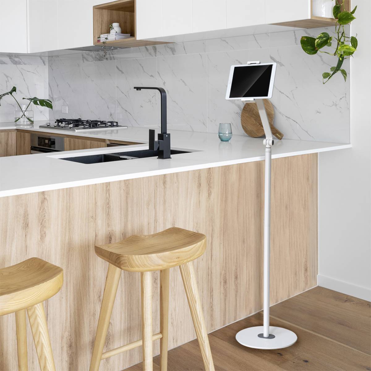 Cucina moderna con piano di lavoro in legno, due sgabelli e un supporto per tablet. Armadi bianchi, schienale in marmo e piante come decorazione.