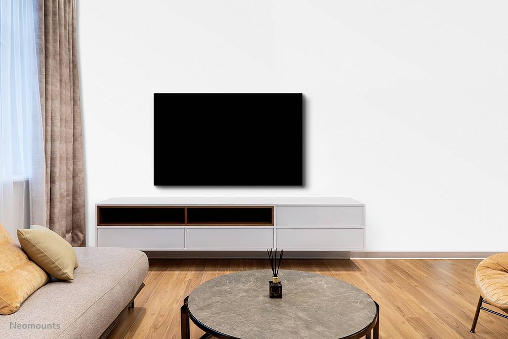 Soggiorno con divano grigio, tavolino rotondo e grande televisore nero su parete bianca. Pavimento in parquet chiaro e finestra a tutta altezza con tenda.