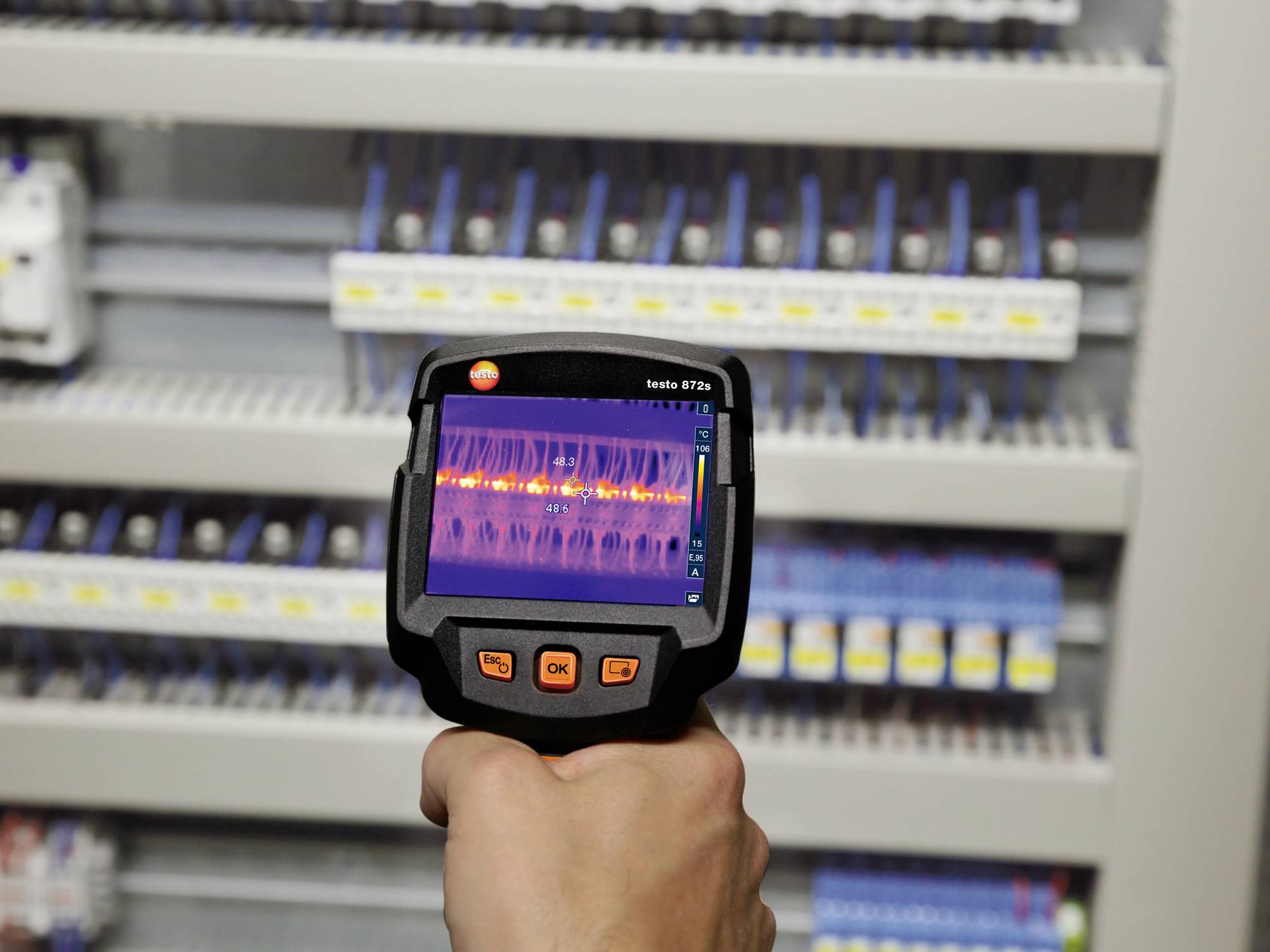 Una mano tiene una termocamera davanti a un quadro elettrico. Lo schermo mostra diverse aree di temperatura nei conduttori.