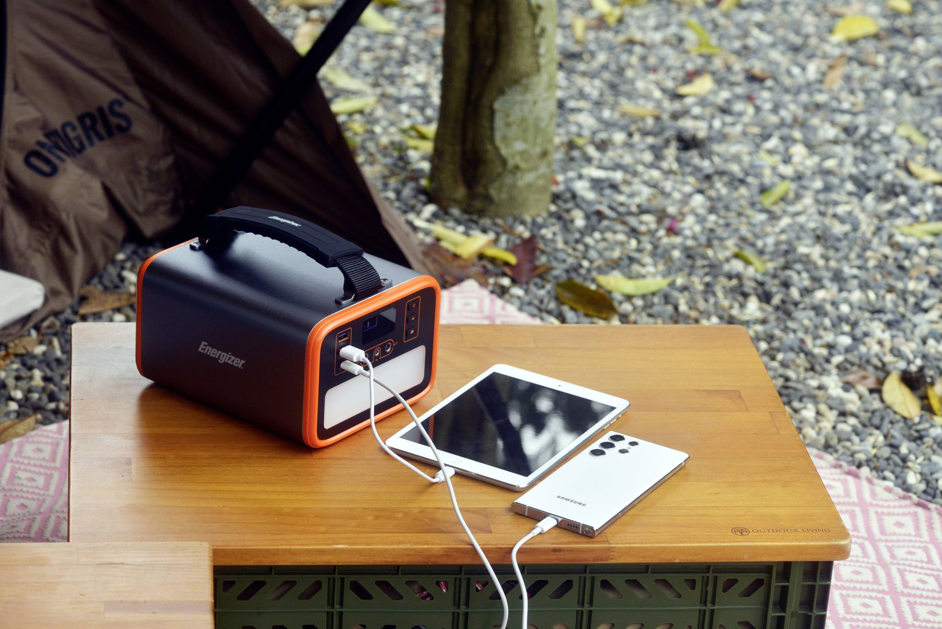 Un power bank è posizionato su un tavolo all'aperto mentre carica uno smartphone e un tablet, circondato da ciottoli e foglie.