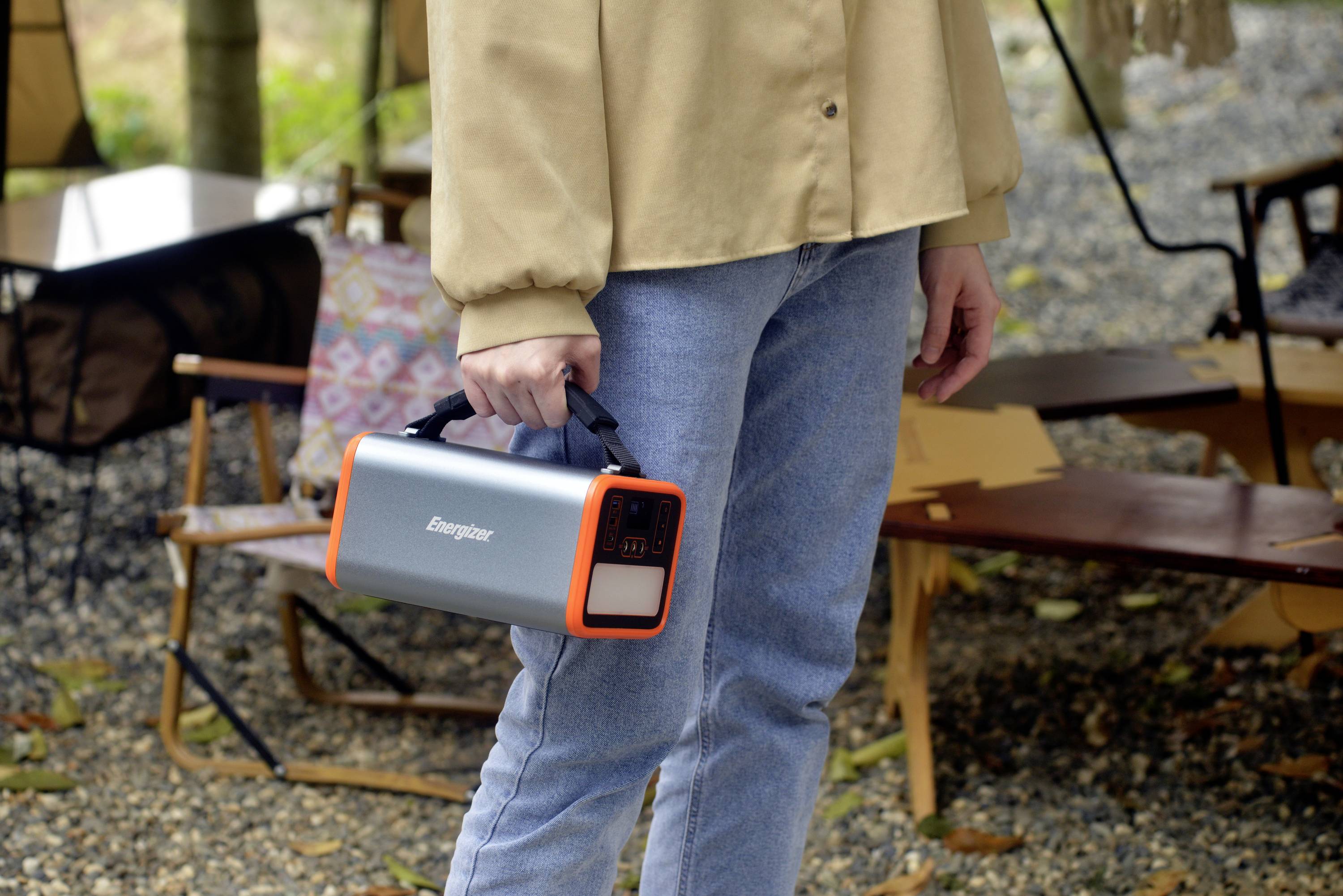 Una persona tiene un altoparlante portatile rettangolare con bordi arancioni in un ambiente esterno da campeggio.