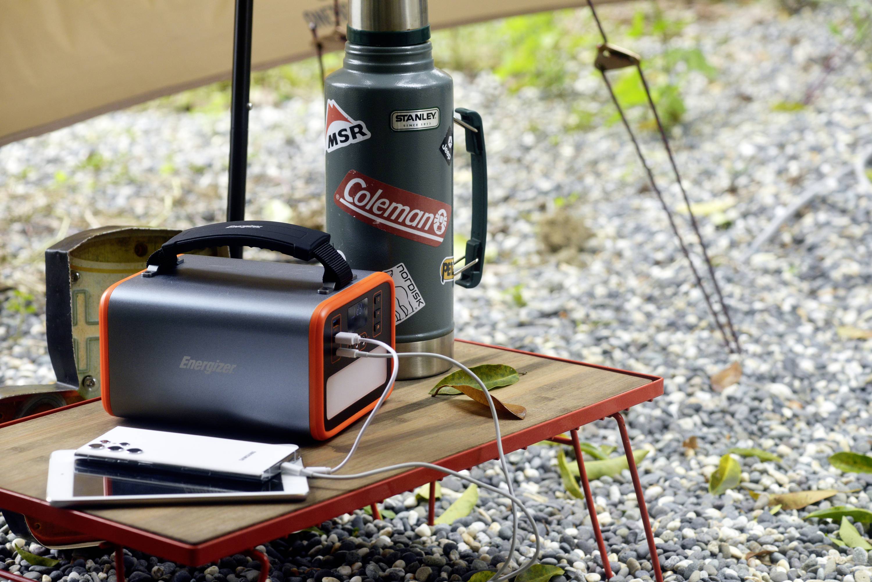 Un caricabatterie portatile sta caricando uno smartphone su un tavolo da campeggio, accanto a un thermos con adesivi, all'aperto.