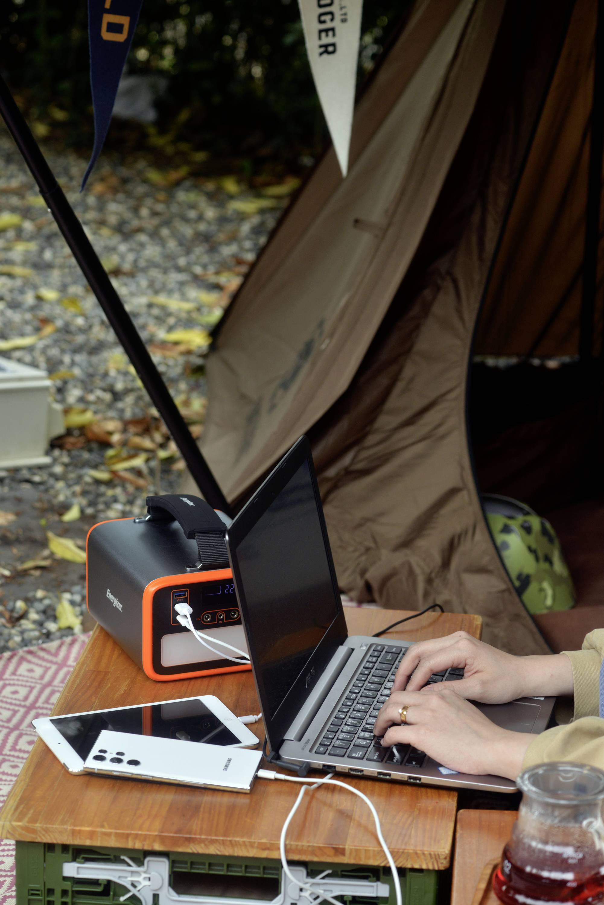 Una persona sta digitando su un laptop davanti a una tenda aperta. Un caricabatterie portatile e un tablet sono posizionati accanto sul tavolo.