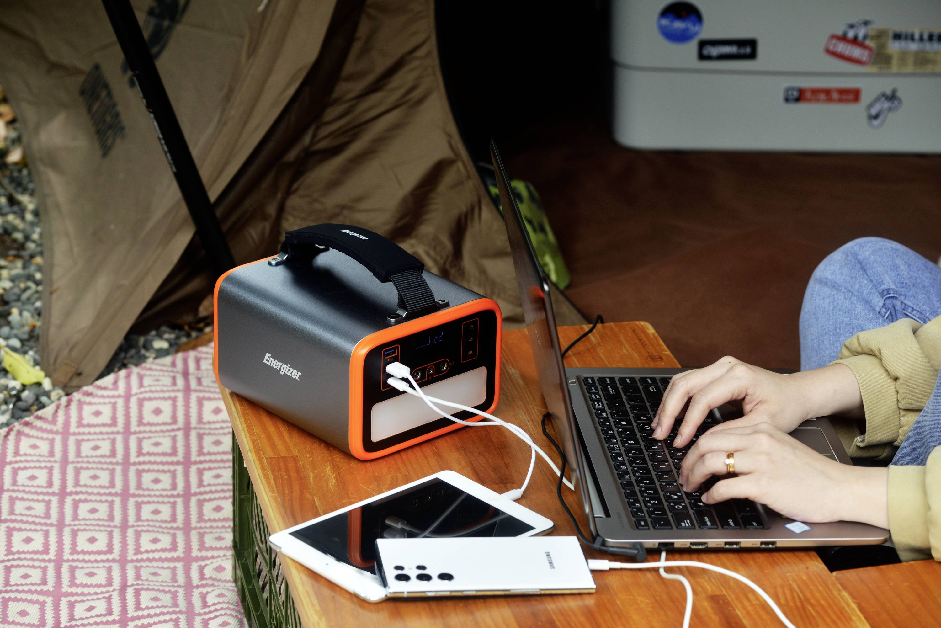 Una persona lavora su un laptop collegato a un caricabatterie portatile. Un tablet è posizionato accanto. Sullo sfondo si trova una tenda.