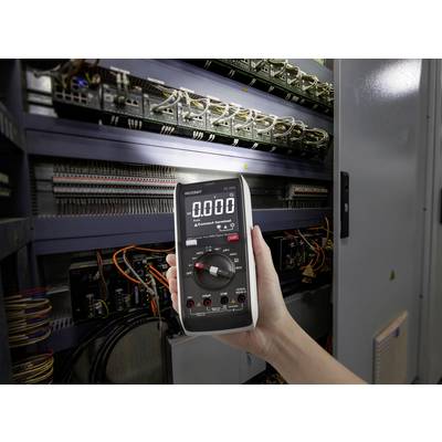 Una mano tiene un multimetro digitale impostato su 0.000 davanti a un quadro elettrico aperto pieno di cavi e componenti elettronici.