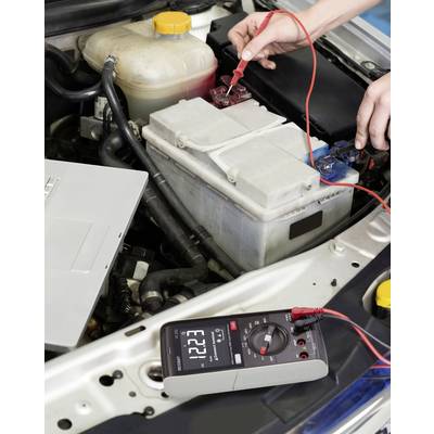 Qualcuno misura la tensione di una batteria dell'auto nel vano motore. Il display mostra '12,21'.