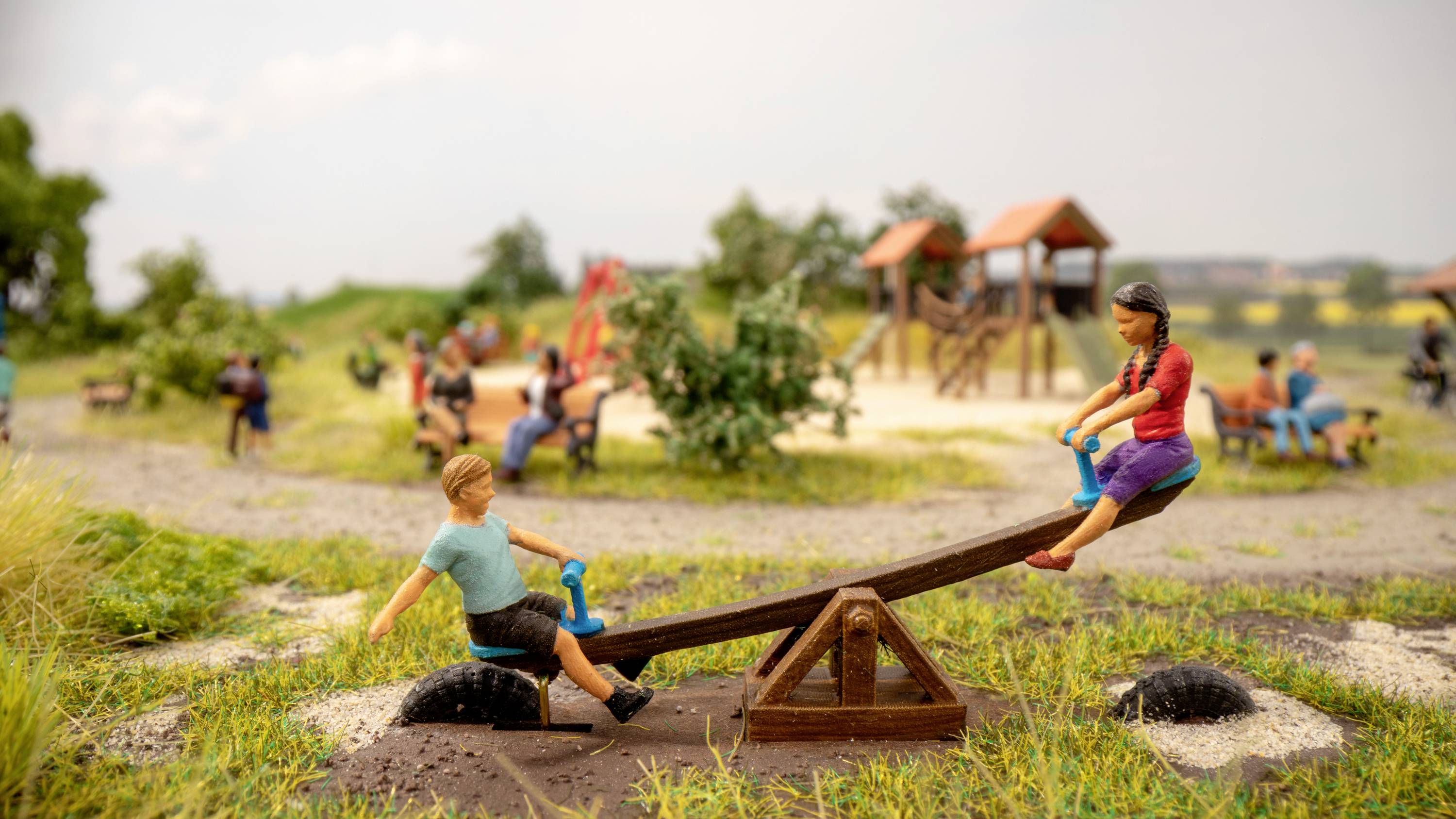 Un modellino in miniatura di un parco giochi mostra due bambini seduti su un'altalena a bilanciere, circondati da altre attrezzature per il gioco e alberi.