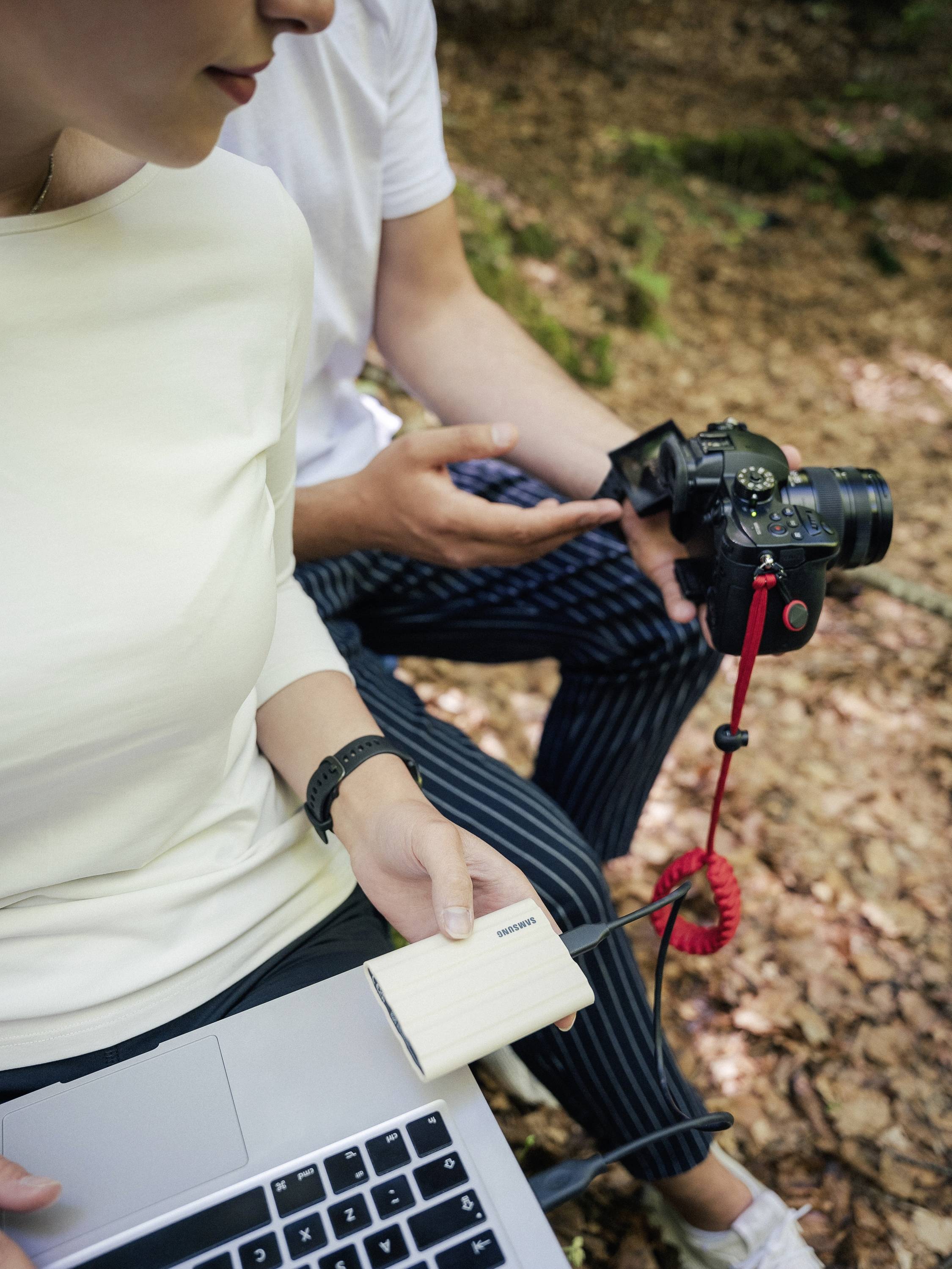 Una persona tiene una fotocamera, mentre un'altra persona trasferisce dati da un dispositivo di archiviazione a un laptop.