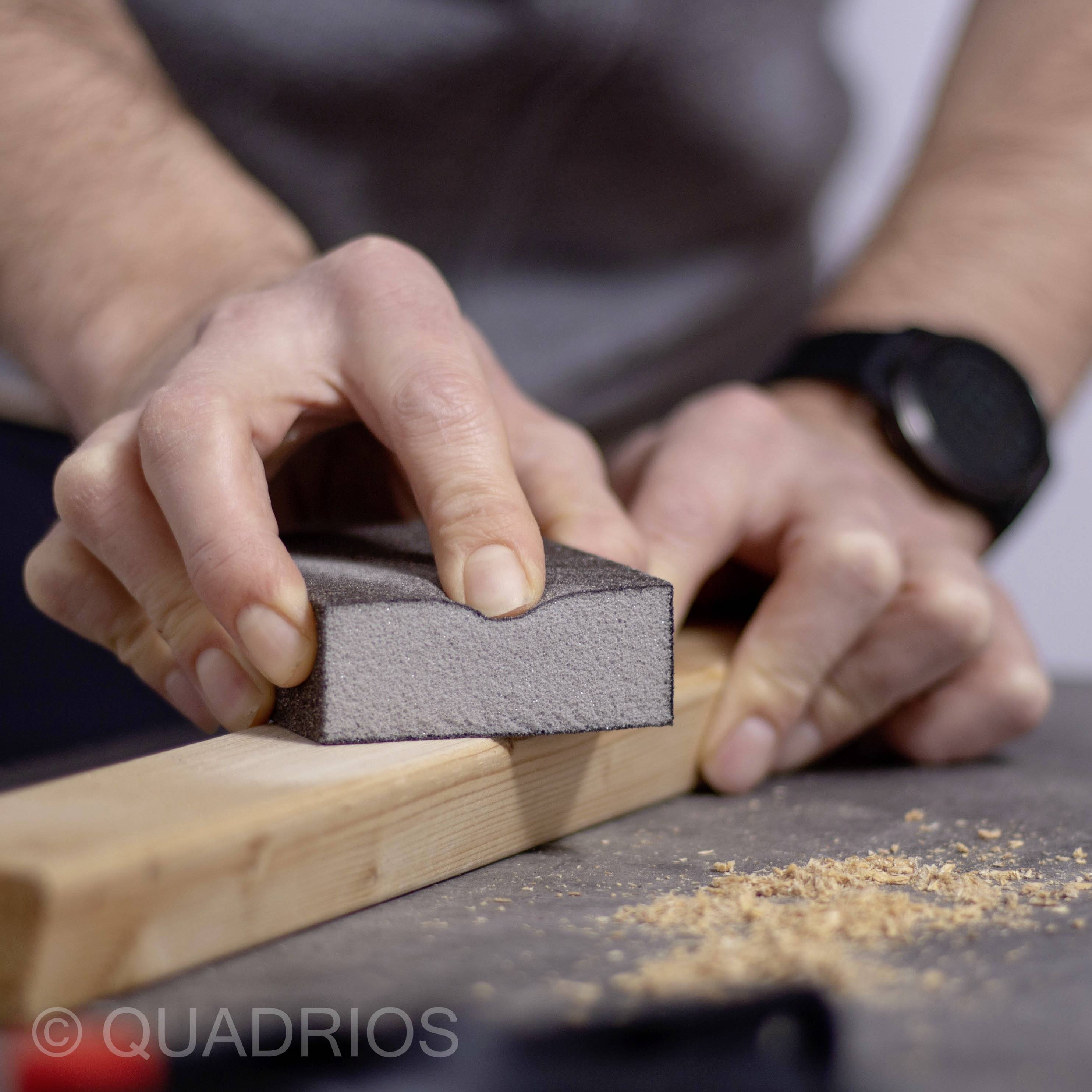 Una persona sta levigando un pezzo di legno con un blocco di carta vetrata, producendo trucioli di legno su una superficie grigia.